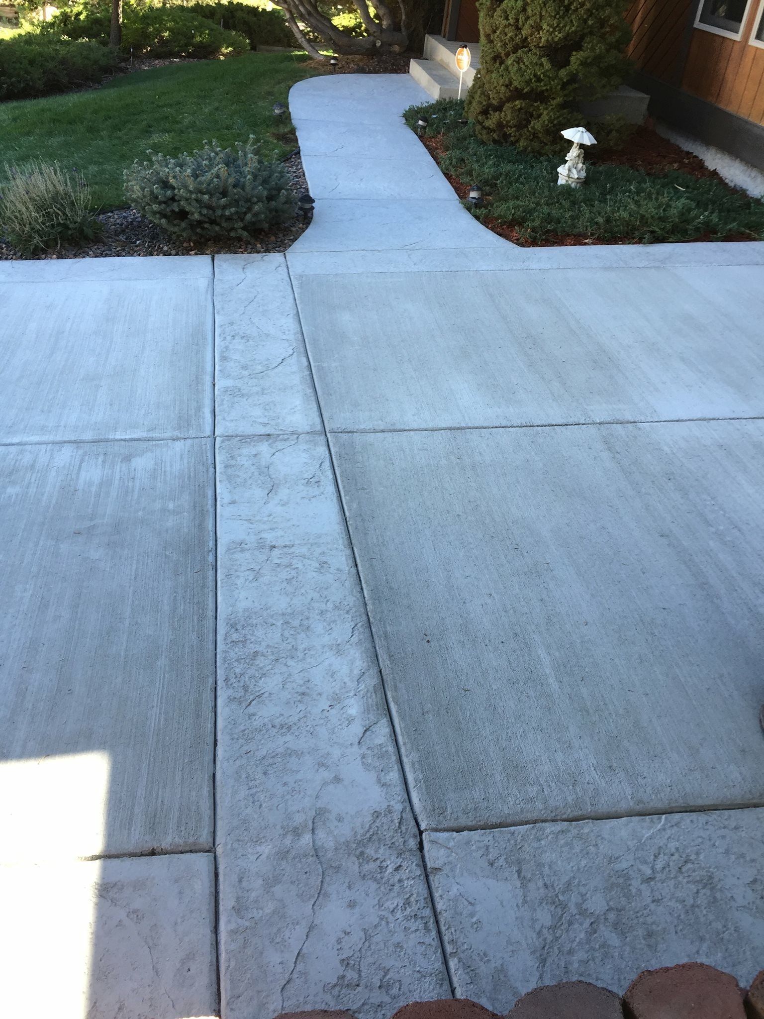 Concrete driveway leading to a sidewalk, bordered by shrubs and a small lawn ornament.