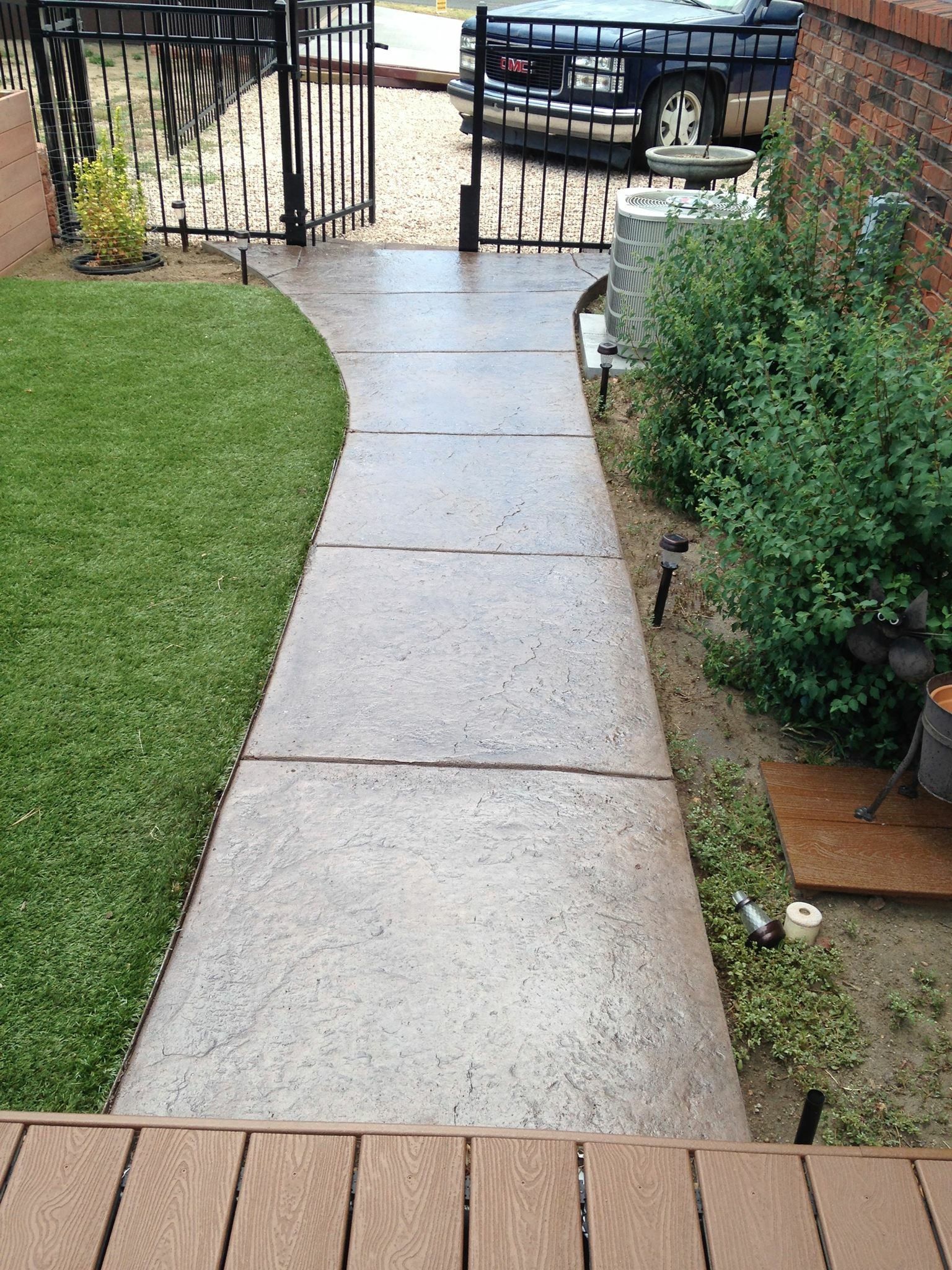 Front yard concrete path leading to a metal gate, bordered by grass and shrubs, with a parked car beyond.