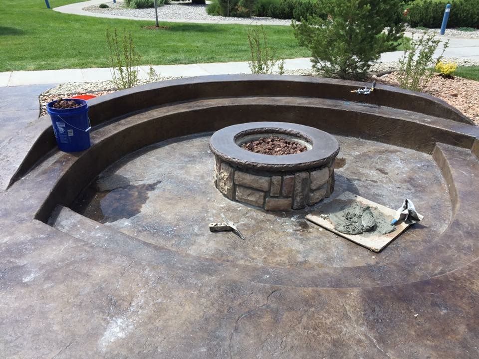 Curved concrete fire pit with stone ring and tools on a patio in a landscaped yard