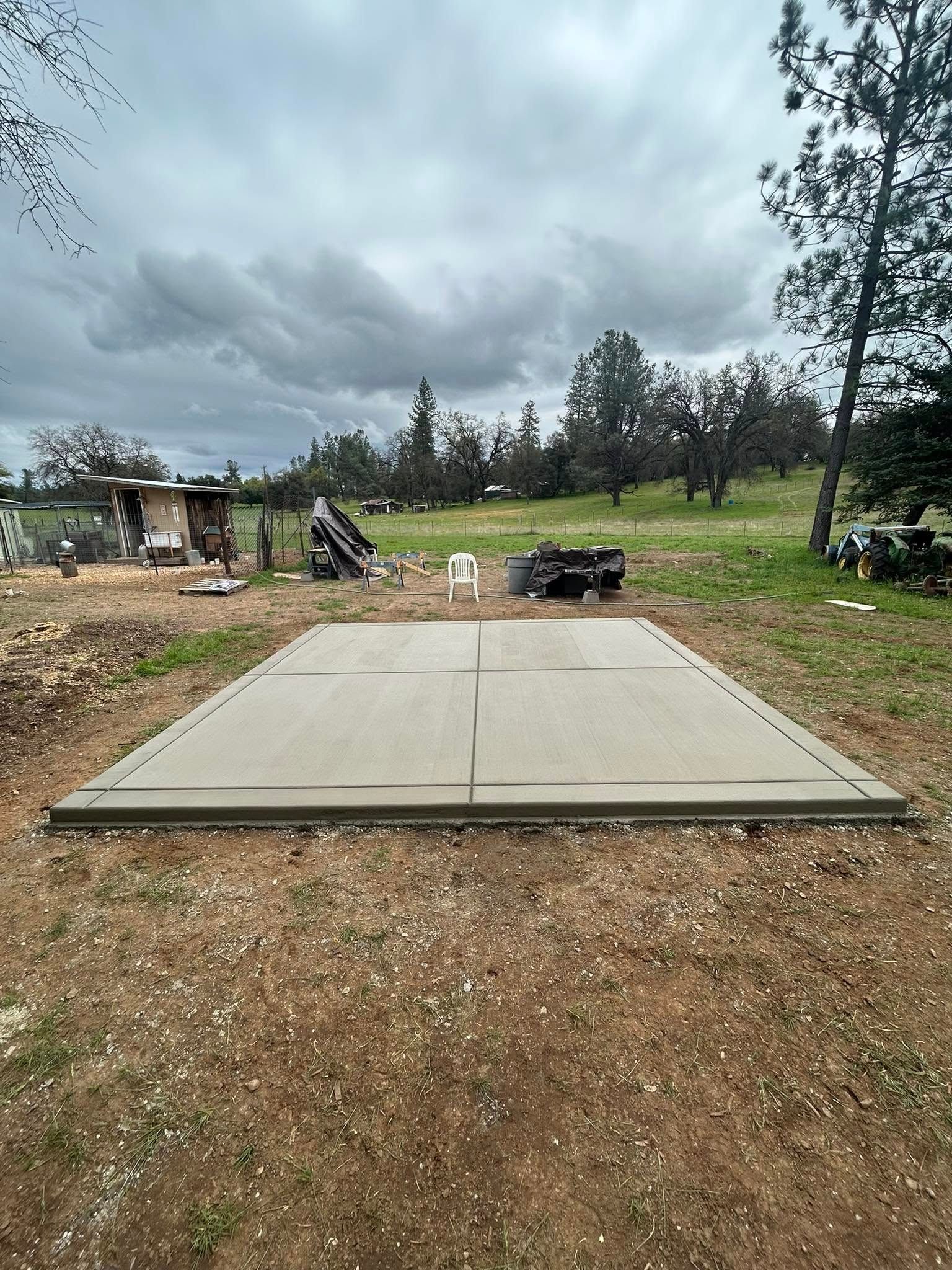 Concrete pad in a rural yard with scattered debris, trees, and cloudy sky.