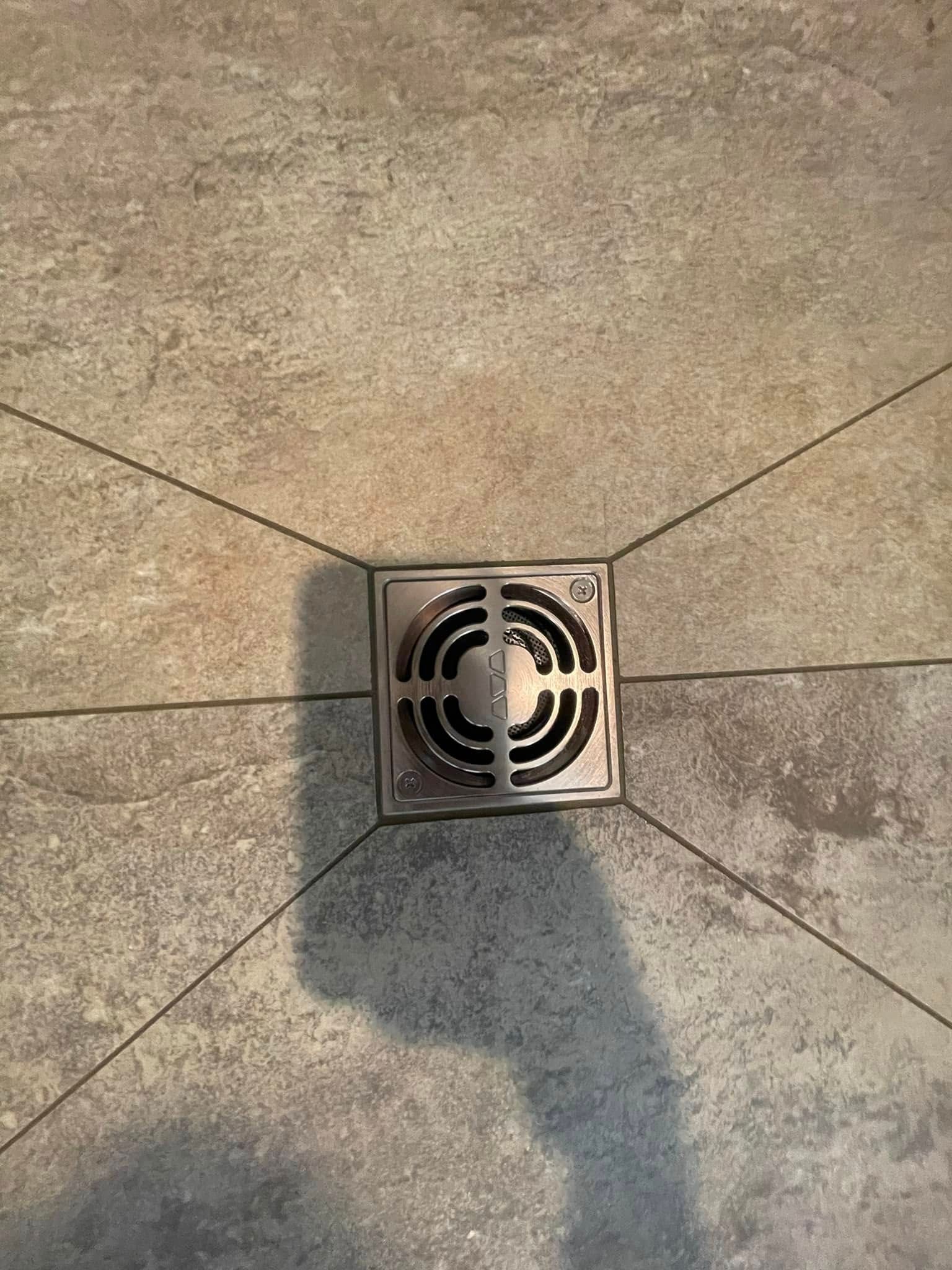 Square shower drain in tiled floor, grout lines radiating outward.