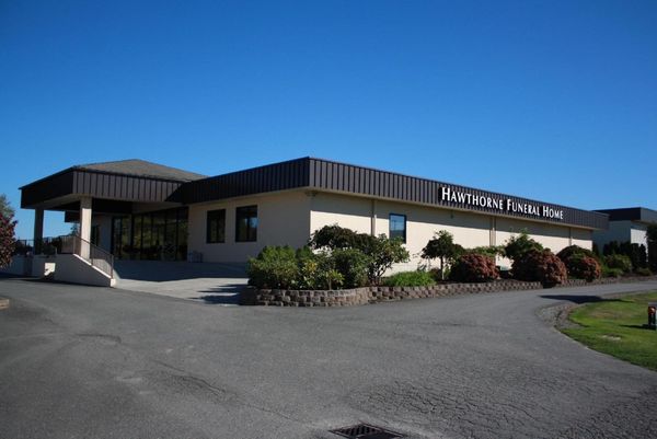A low, gray building with American flag, sign, and driveway. Landscaped garden. Bright day.
