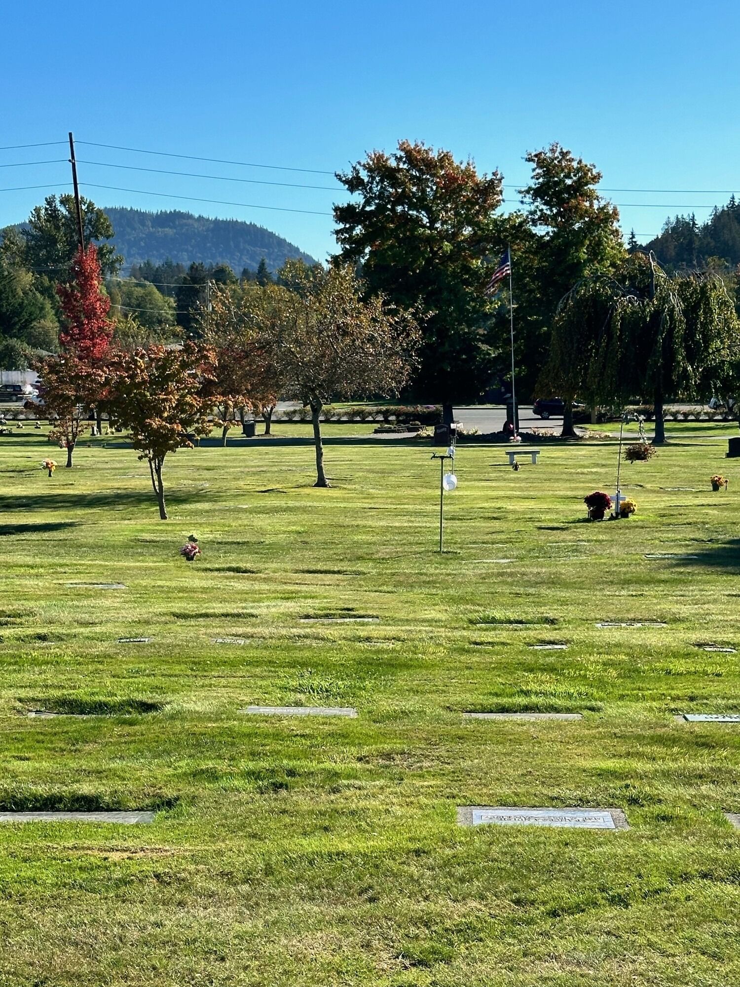Lush green cemetery with trees, some showing fall colors. Mountains and blue sky in the background.