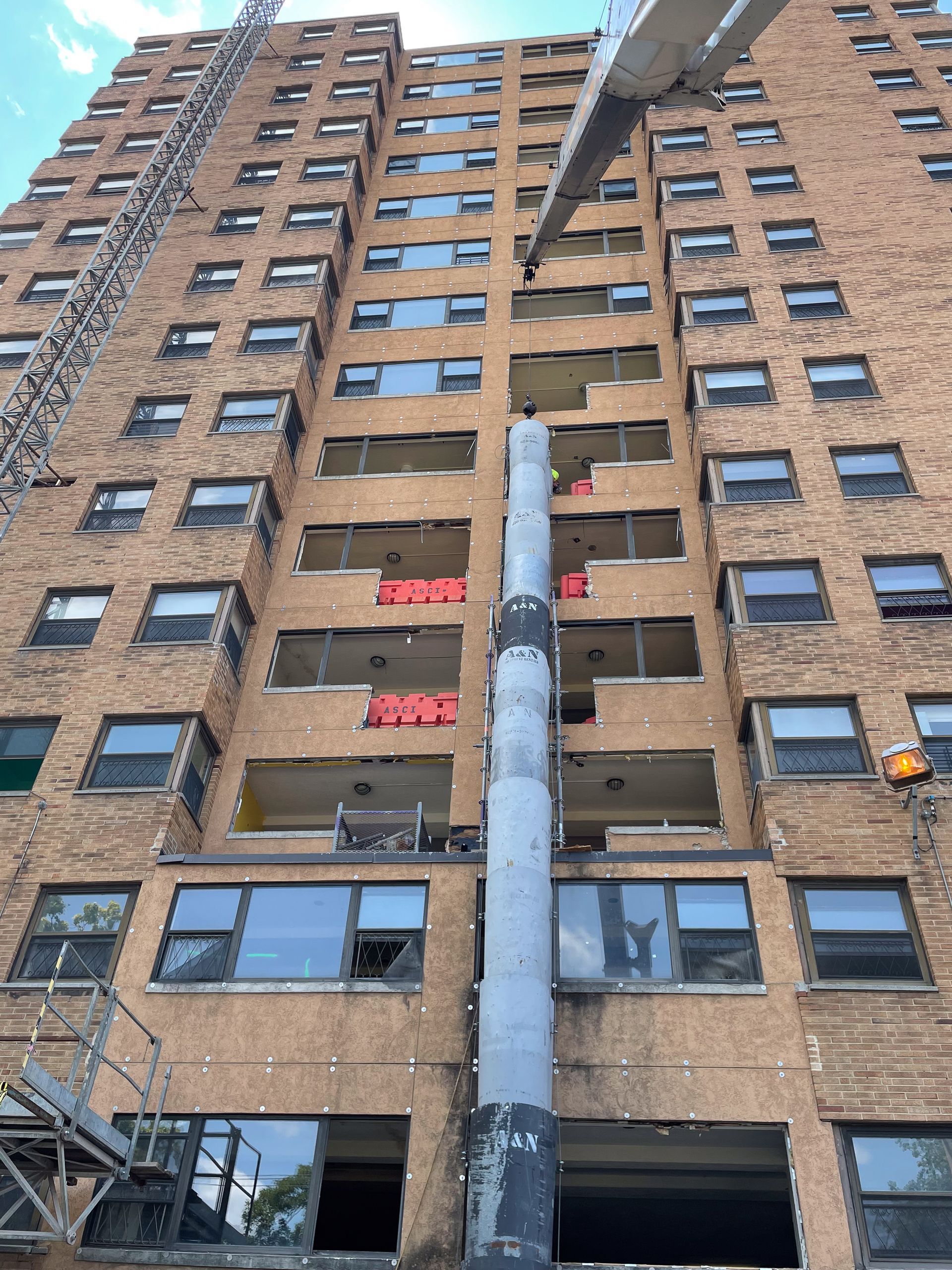 A crane from A&N Crane Rental is hanging a metal trash chute on the side of a tall brick building.
