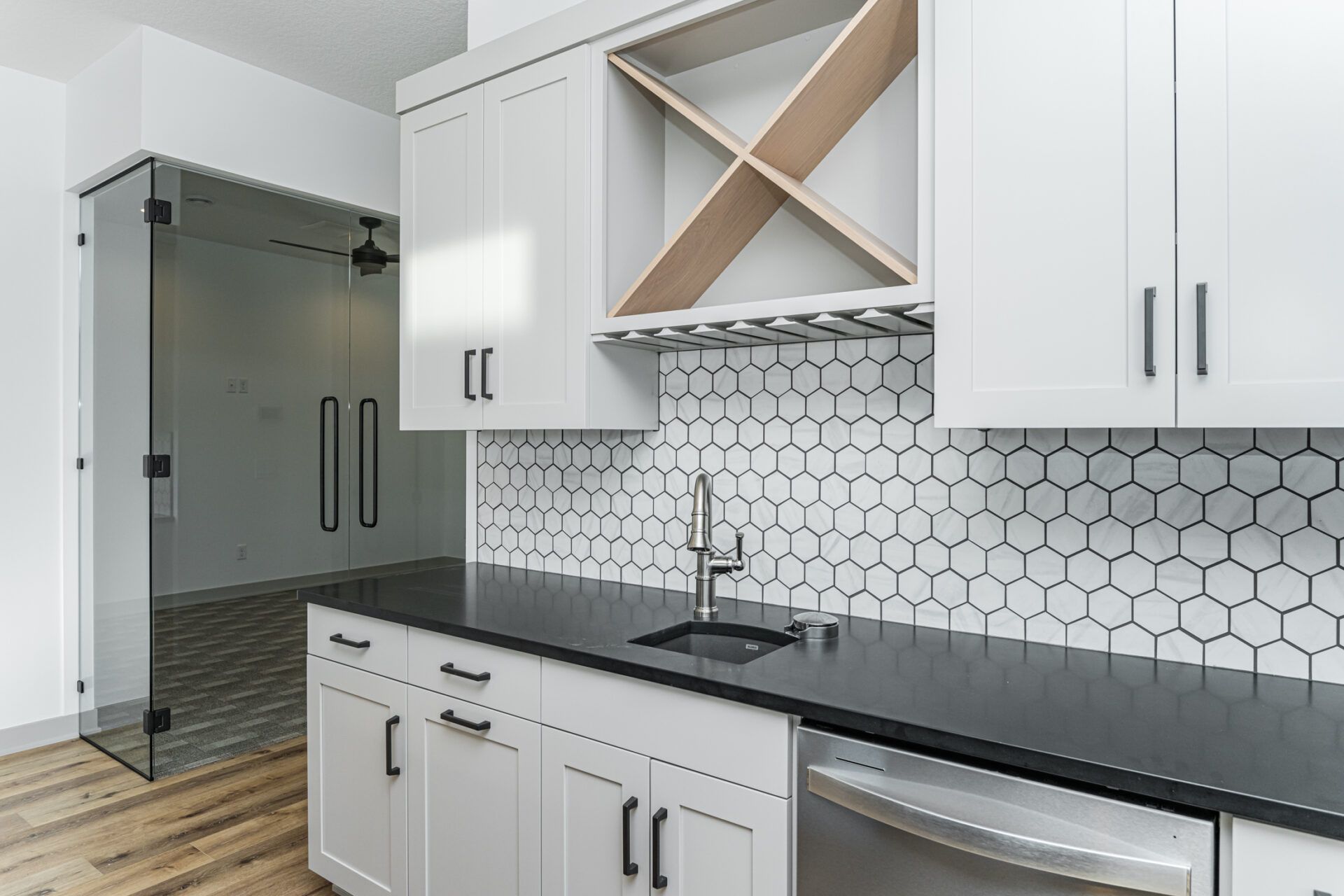 Modern white kitchen with black countertop, hexagon tile backsplash, and glass door to a room.