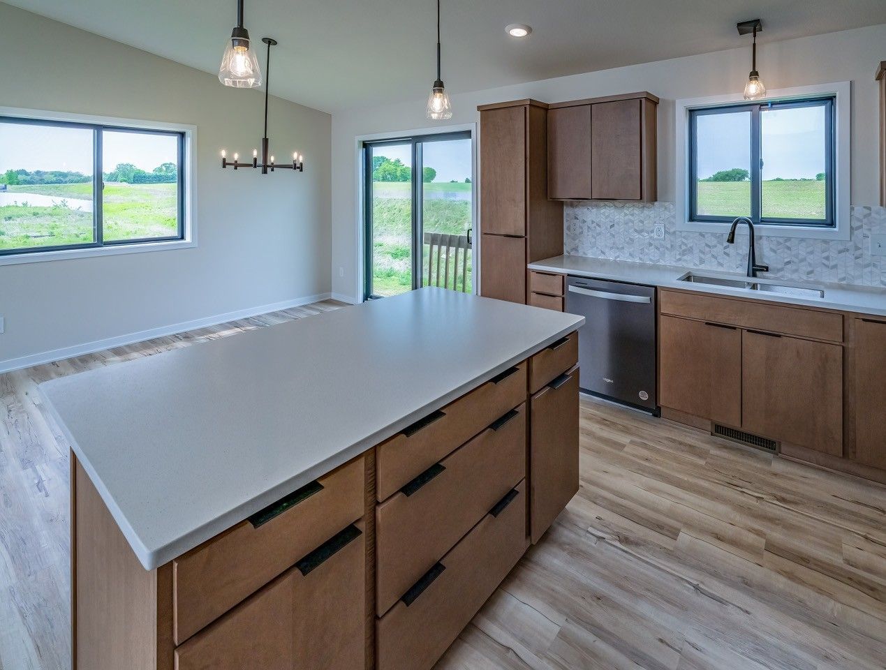 Modern kitchen with island, wooden cabinets, stainless steel appliances, and large windows overlooking a field.