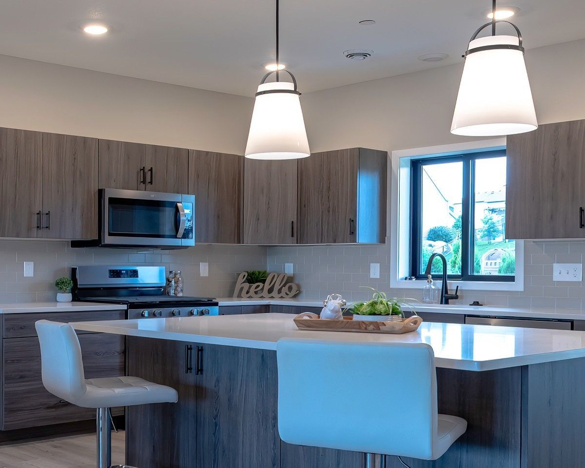 Modern kitchen with island, gray cabinets, stainless steel appliances, pendant lights, and two white chairs.