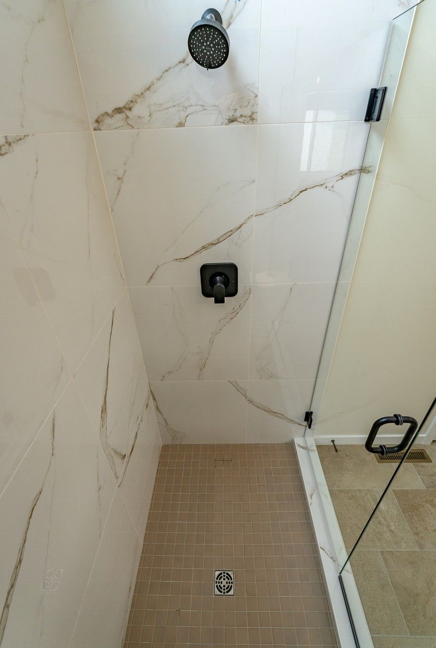 Shower with white marble-like tile walls, beige floor, black shower fixtures, and glass door.