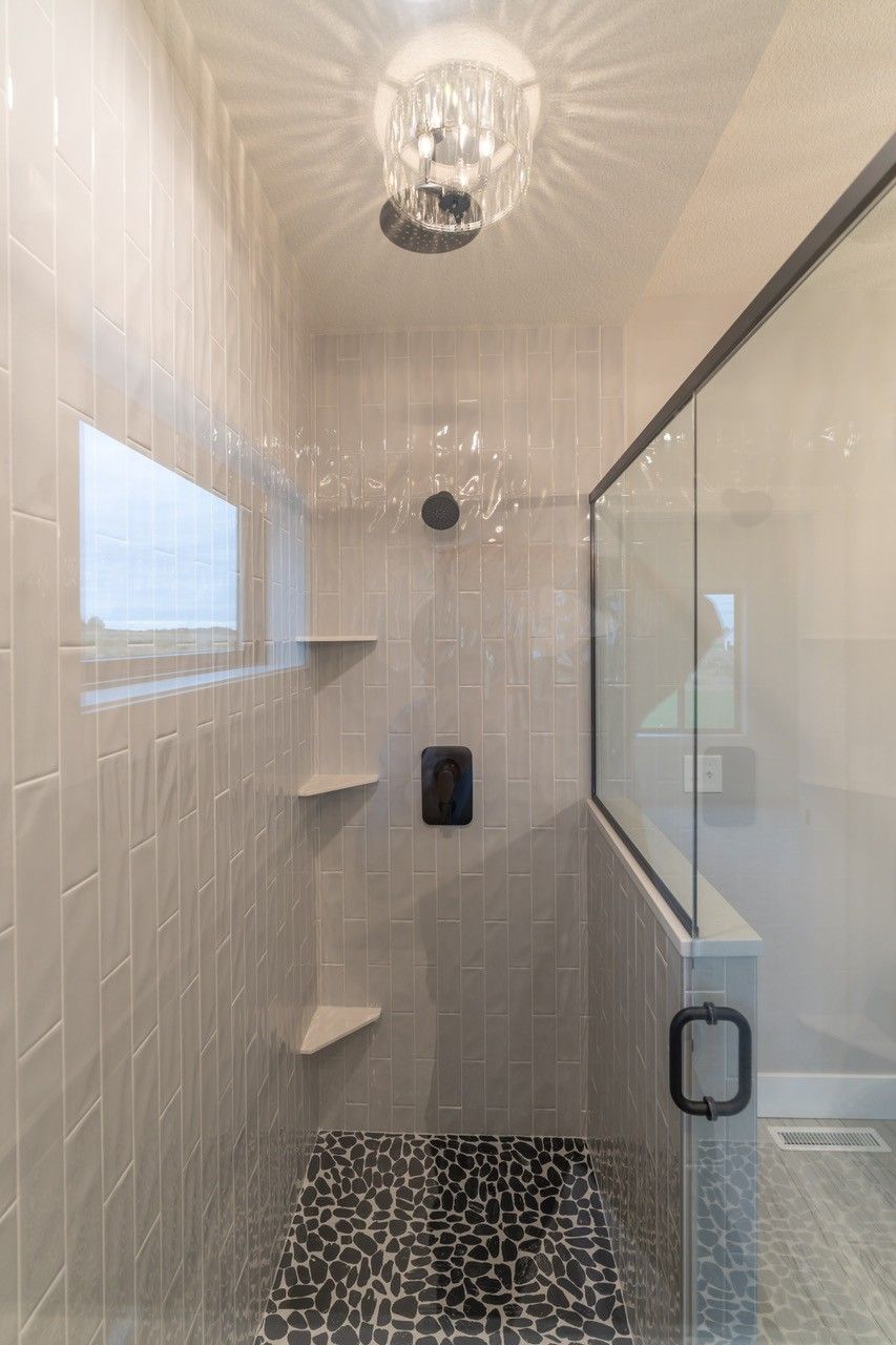 Shower with gray tile walls, pebble floor, and glass enclosure. A crystal chandelier hangs overhead.
