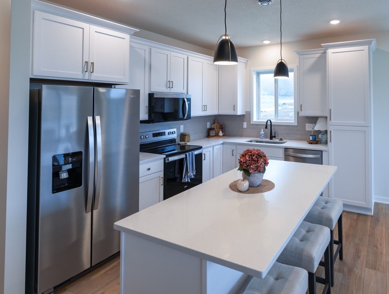 White kitchen with island, stainless steel refrigerator, and hanging lights.