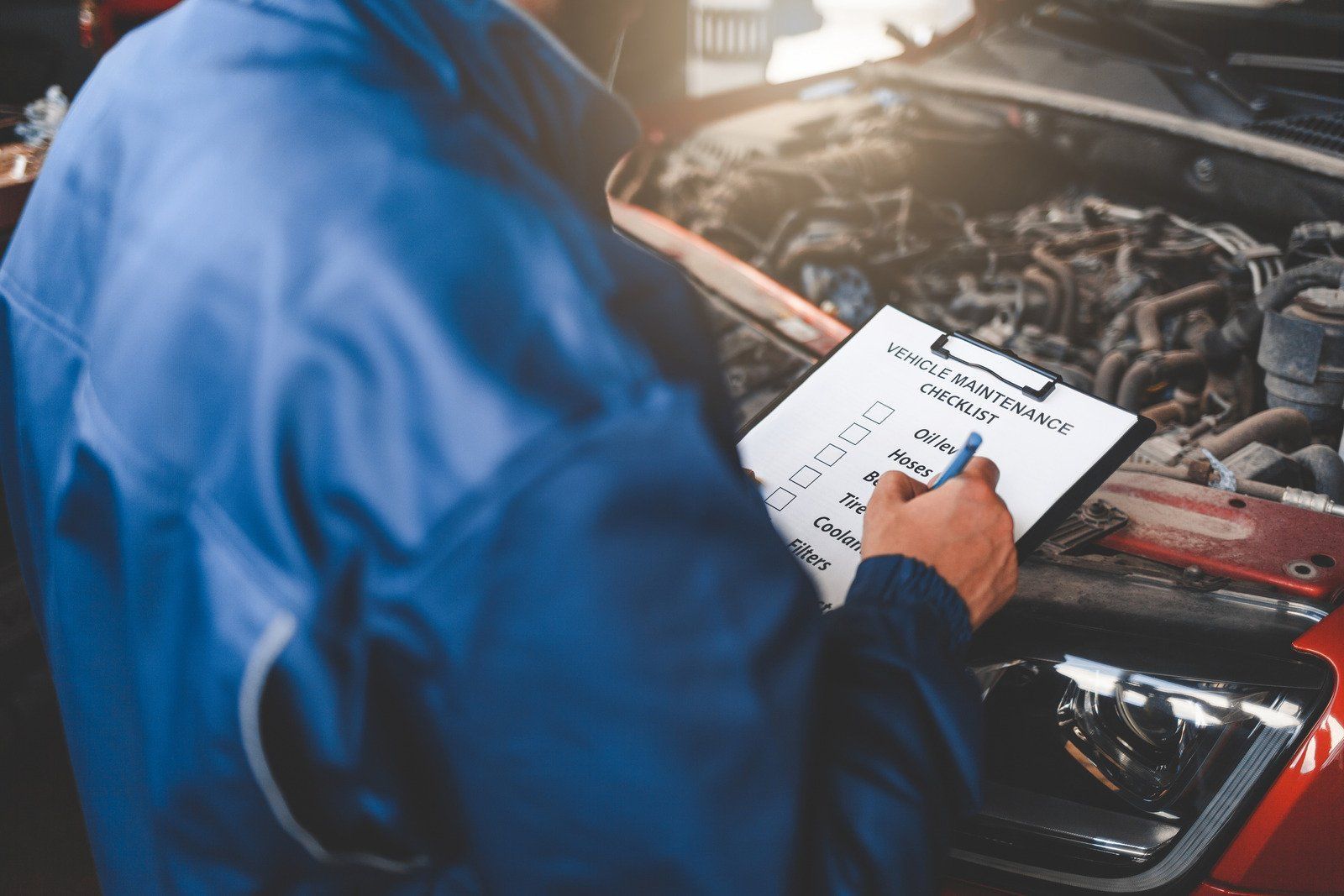 A mechanic is writing on a clipboard while looking under the hood of a car.