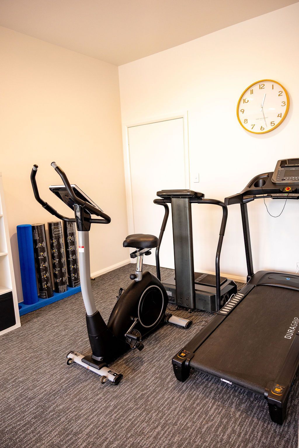 Home exercise room with treadmill, stationary bike, and elliptical on carpeted floor near a wall clock. — Forerunner Physio In Taree, NSW