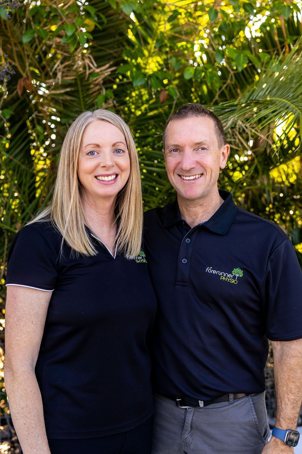 Two people in black shirts smiling outdoors in front of green foliage.— Forerunner Physio In Taree, NSW