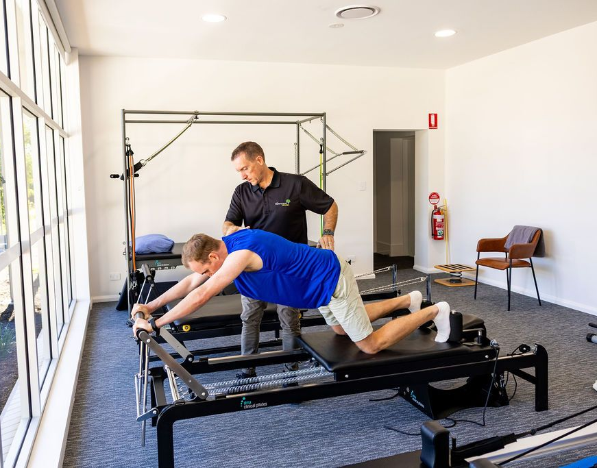 Person exercising on a Pilates reformer while a trainer assists in a bright studio — Forerunner Physio In Taree, NSW