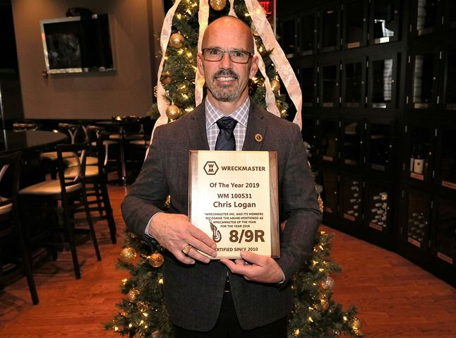 A man in a suit smiles while holding an award plaque in front of a decorated Christmas tree in an indoor restaurant.