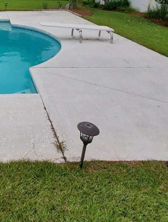 A concrete patio with a swimming pool in the background and a lamp in the foreground.