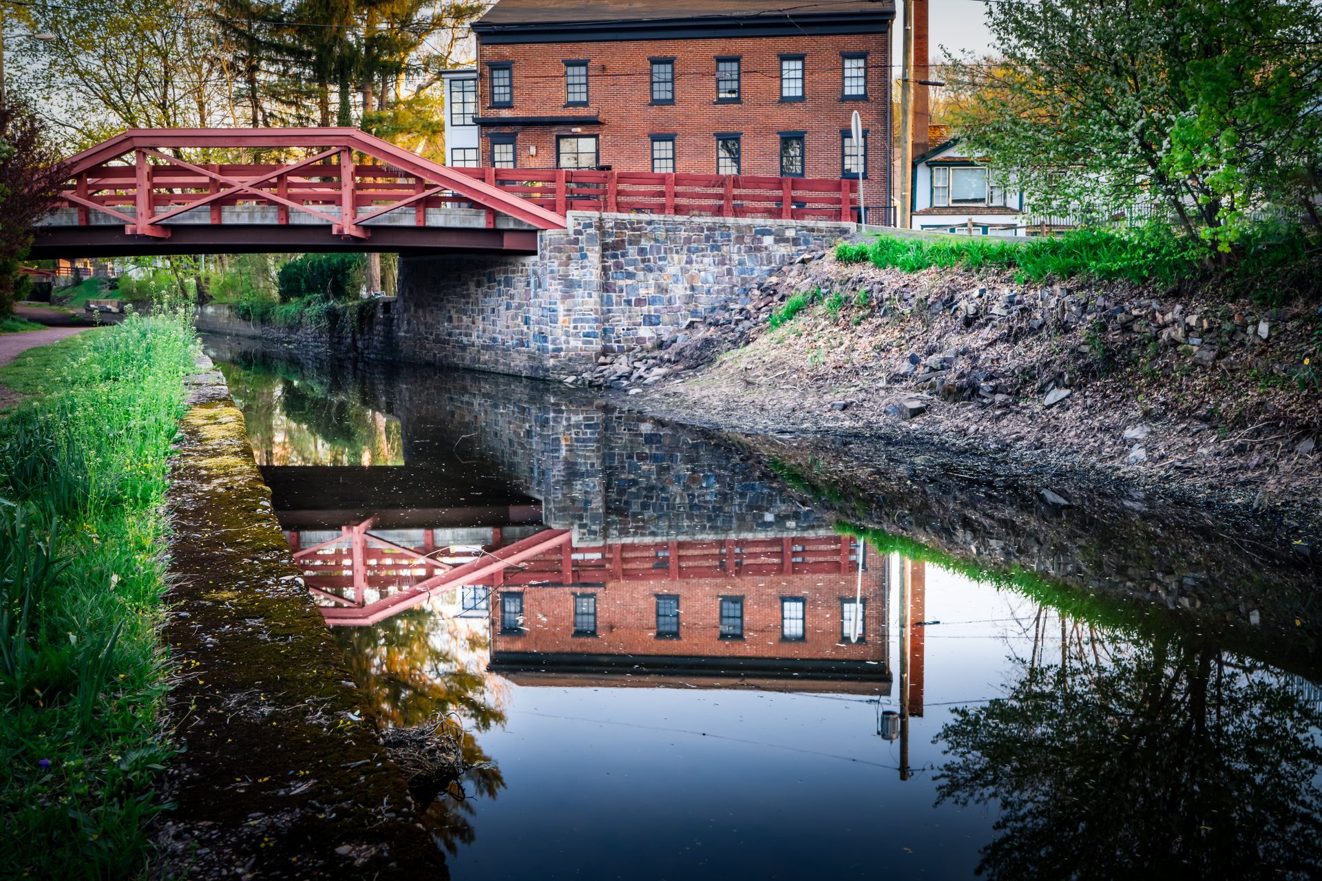Brick building reflected in canal water beneath a red bridge. Green grass and trees line the canal. Brick building reflected in canal water beneath a red bridge. Green grass and trees line the canal.