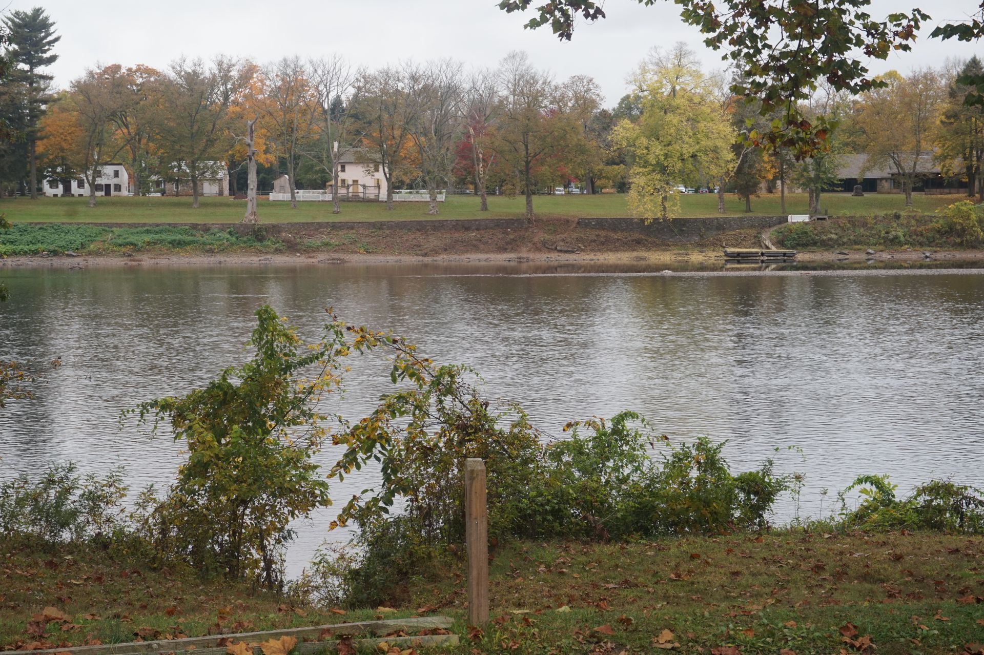 A calm lake reflects trees with autumn colors. Houses line the far bank under an overcast sky. A calm lake reflects trees with autumn colors. Houses line the far bank under an overcast sky.