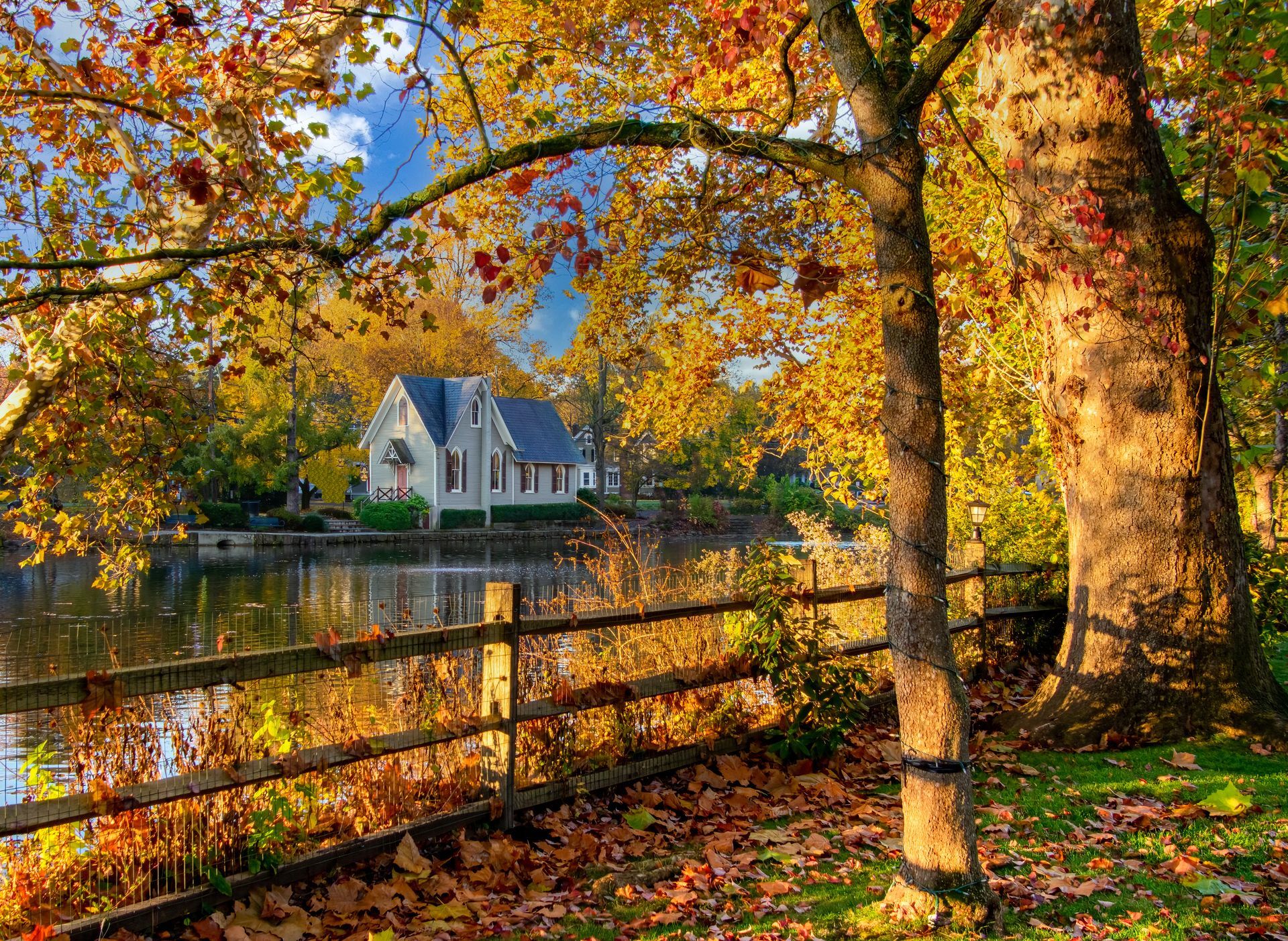 Autumn scene: a house by a lake with a wooden fence. Yellow leaves and trees frame the view under a blue sky. Autumn scene: a house by a lake with a wooden fence. Yellow leaves and trees frame the view under a blue sky.