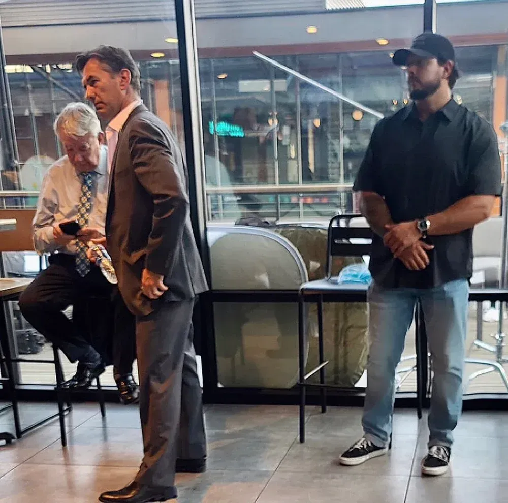 Three men inside a building. One in a suit, one on a phone, the last wearing jeans and a baseball cap.