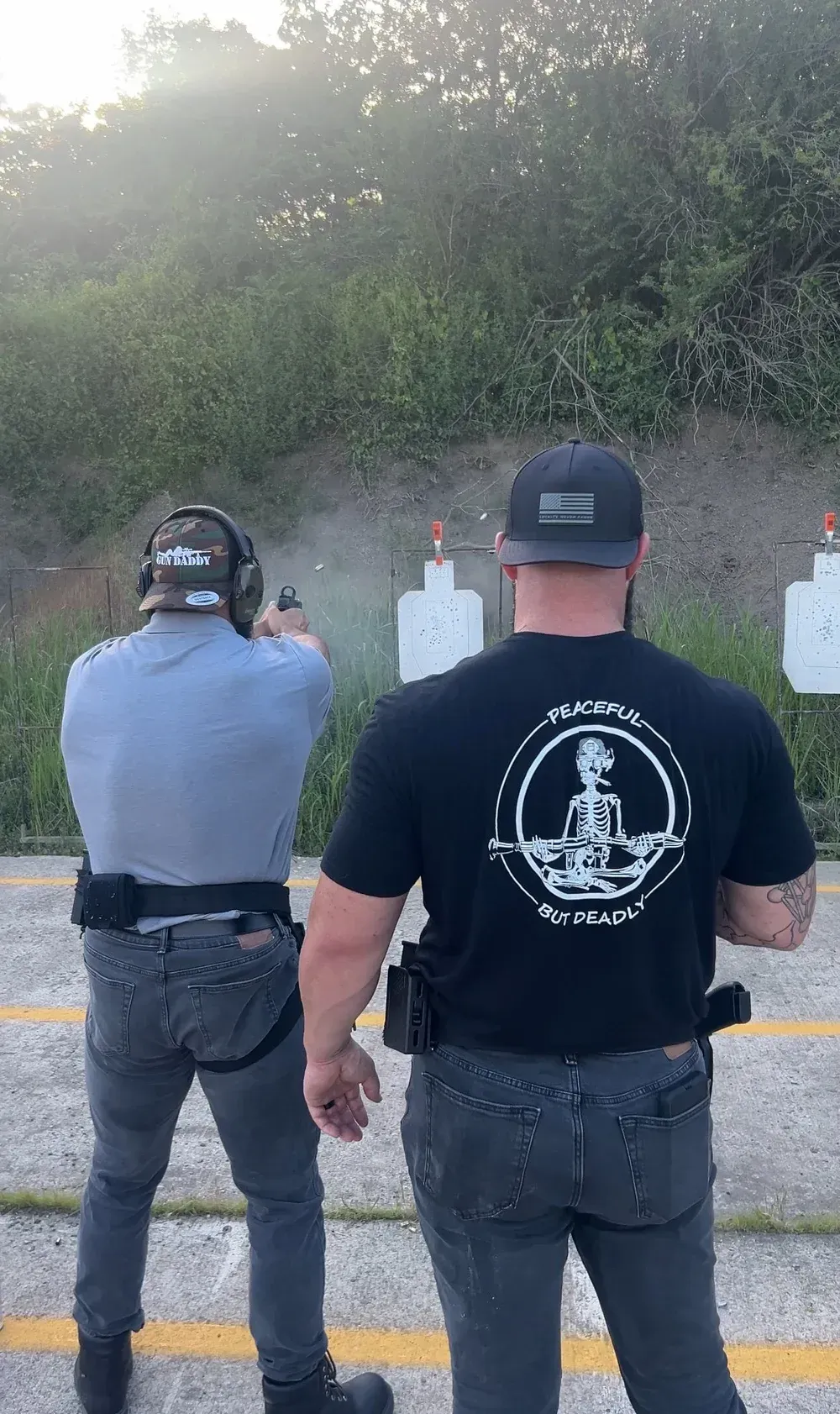 Two men at a shooting range; one fires a handgun. Smoke, targets, and a grassy background.