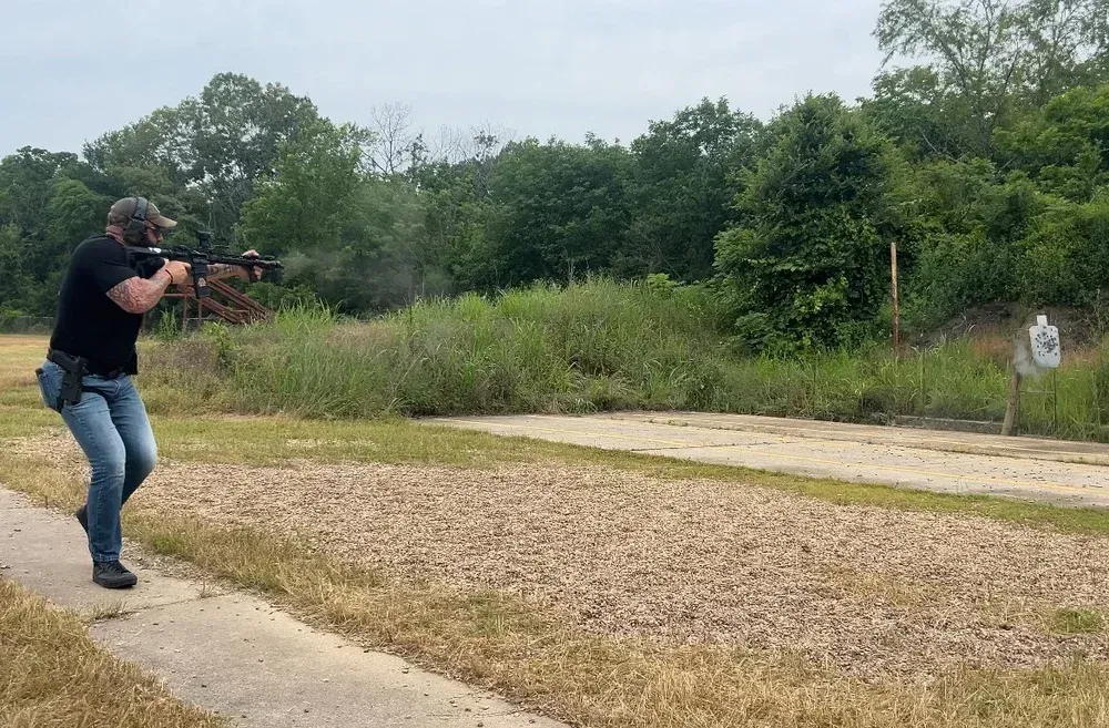 Man in black shirt shoots rifle at outdoor range, target visible.