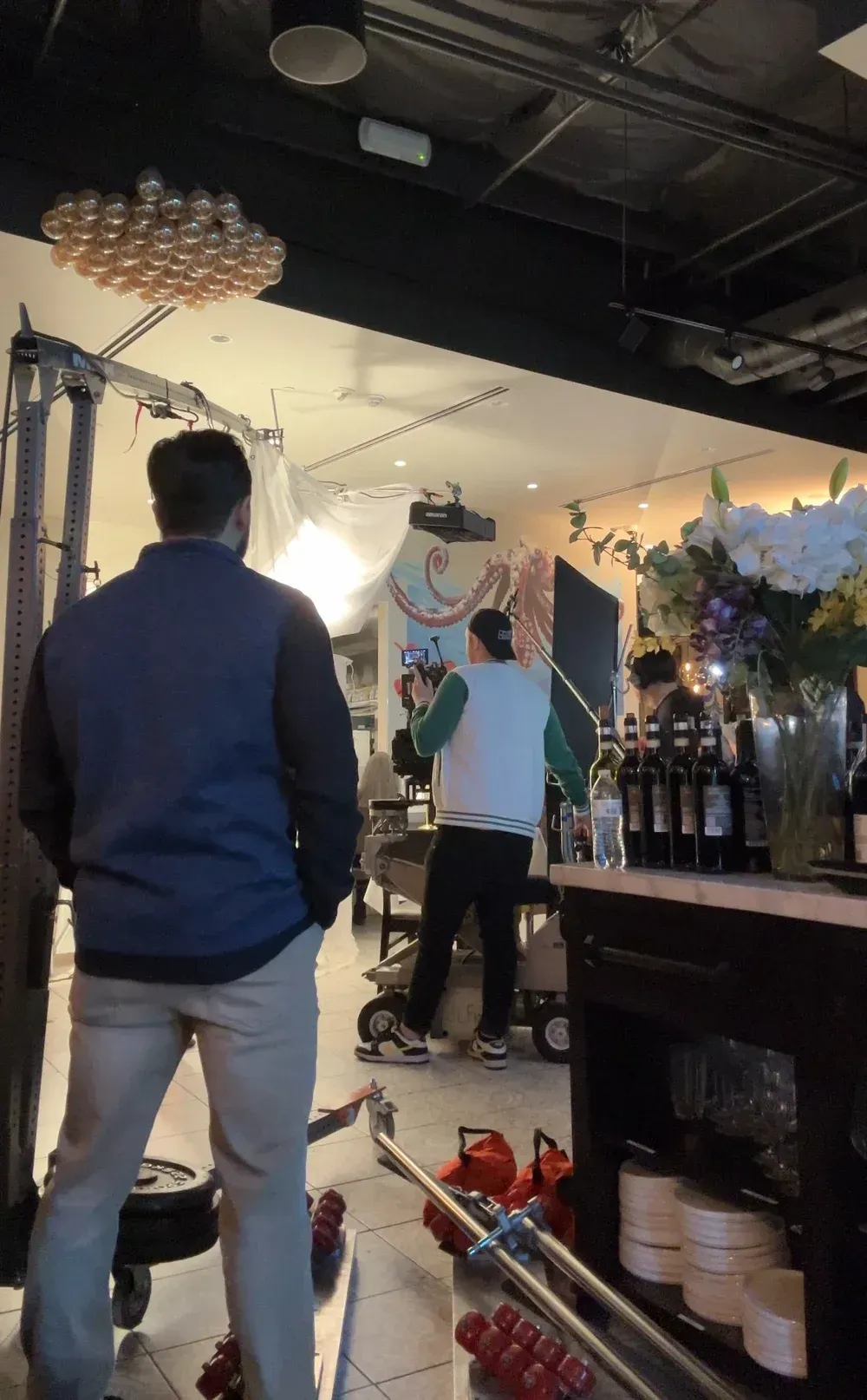 Man in a vest watches filming in a cafe with bar, flowers, and camera equipment visible.