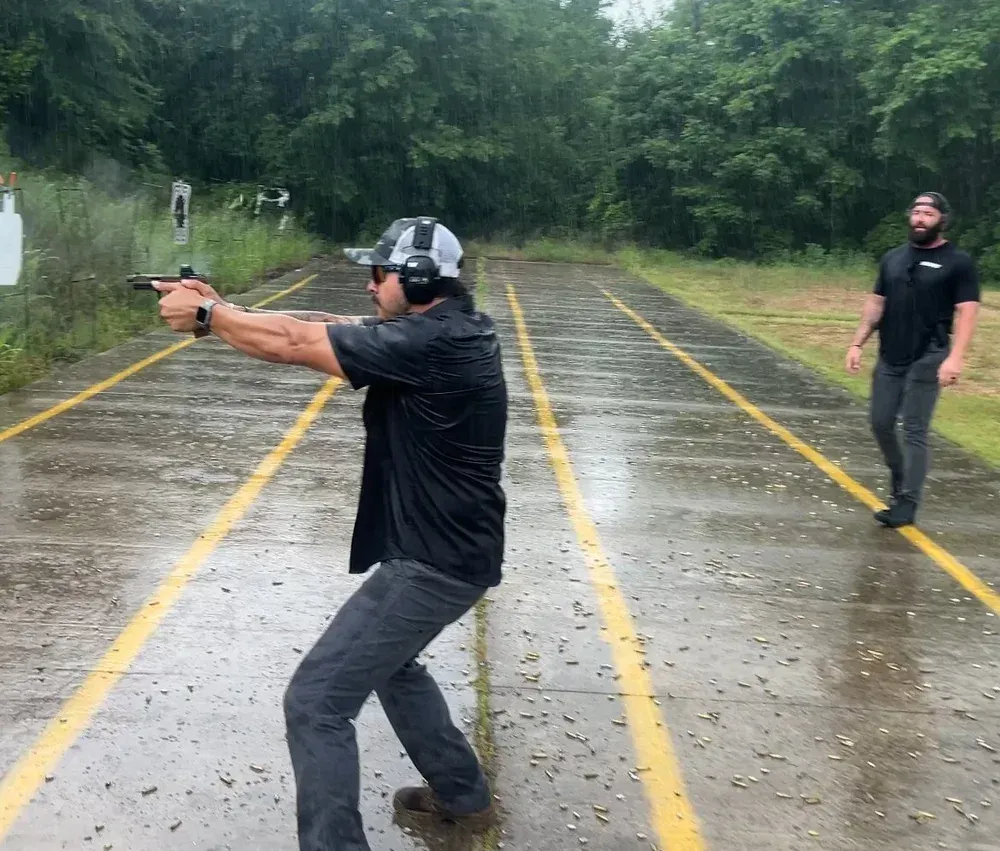 Man shooting a pistol at a target outdoors in the rain; another man watches from behind.