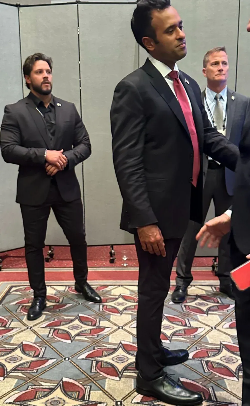 Man in suit with red tie; two other men in suits are standing by him.