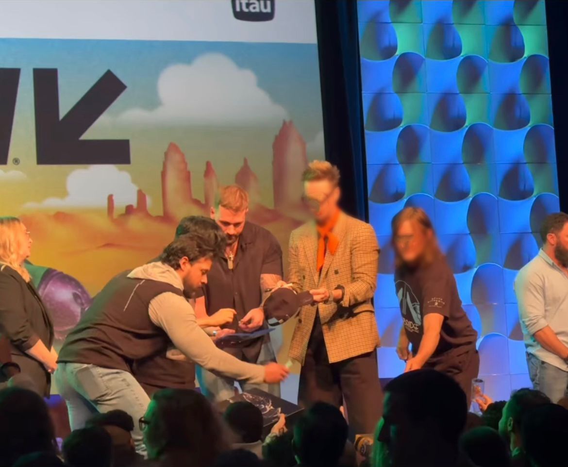 Group on stage; man in orange jacket handing something to another. Event backdrop, audience.