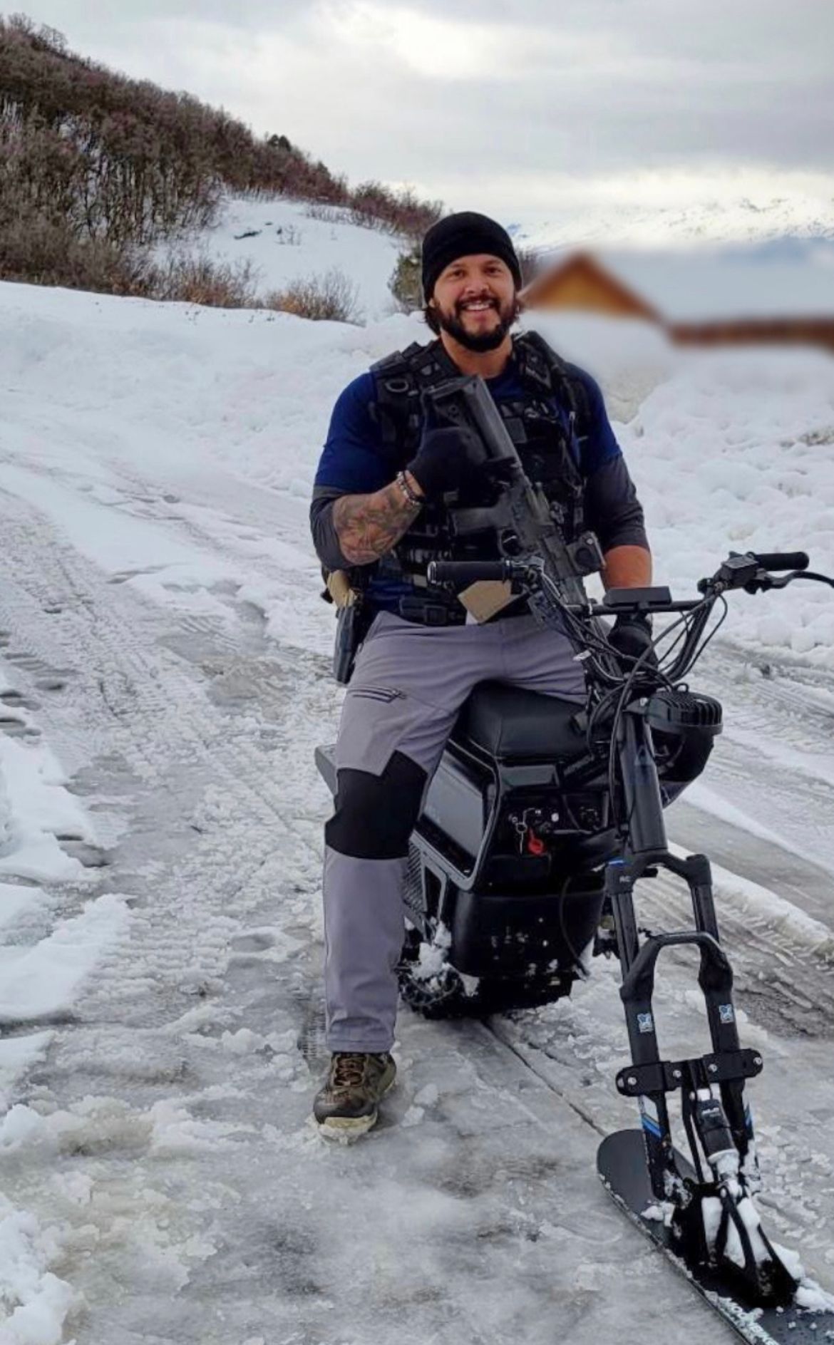 Man on snowbike smiles, holding rifle, in snowy area.
