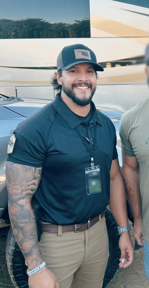 Man in a dark blue polo shirt, khaki pants, and a baseball cap, smiling outdoors. He has tattoos and is wearing an ID badge.