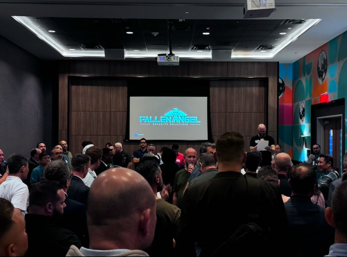 Large group of men in a meeting room, speaker at front, projection screen with company logo, dim lighting.