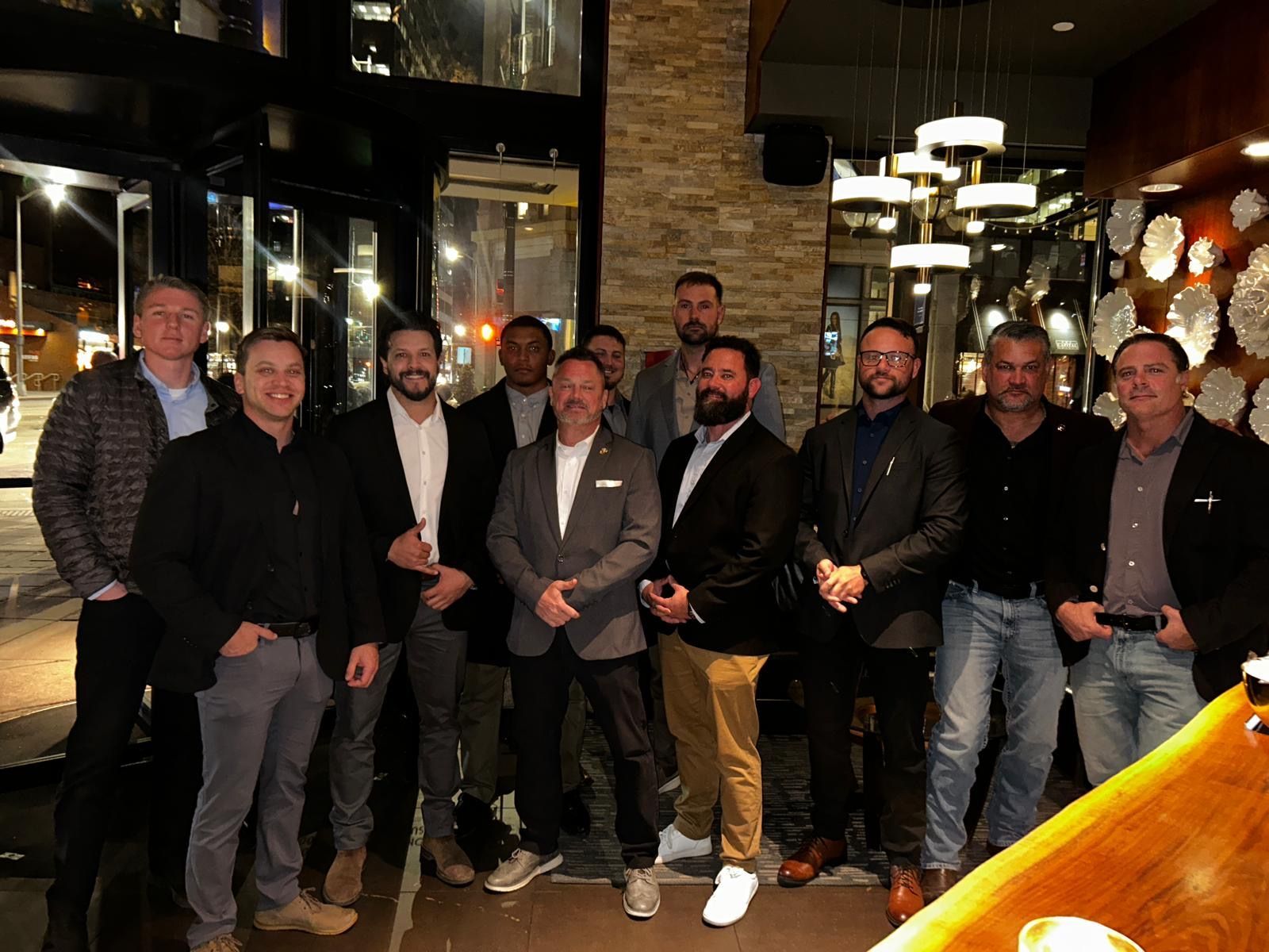 Group of men in business attire, standing inside a restaurant with a dark interior and some outdoor lighting.