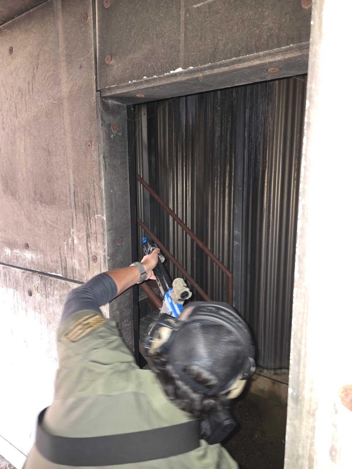 Person in tactical gear aims a weapon at a dark doorway inside a concrete structure.