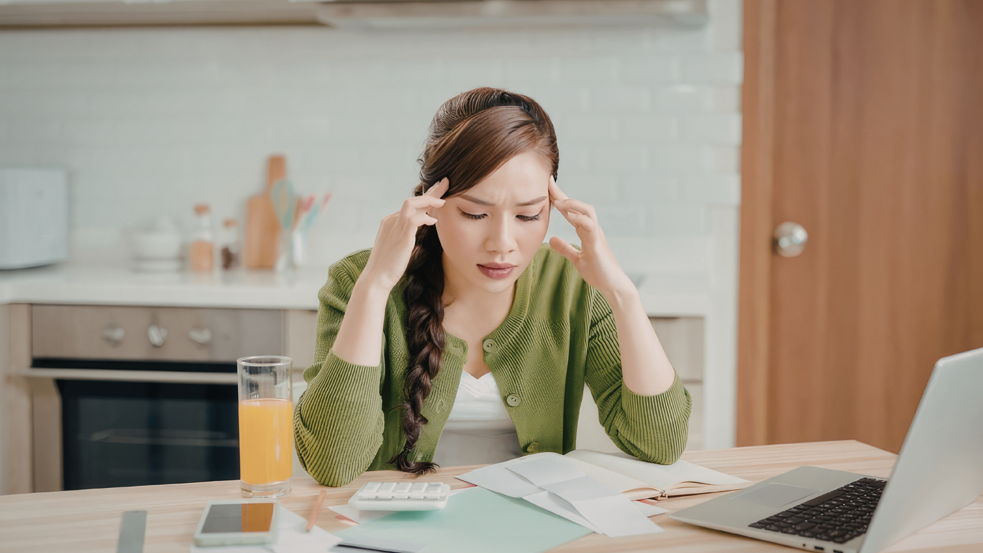 Stressed person holding temples at a kitchen table with papers, laptop, and a glass of juice.
