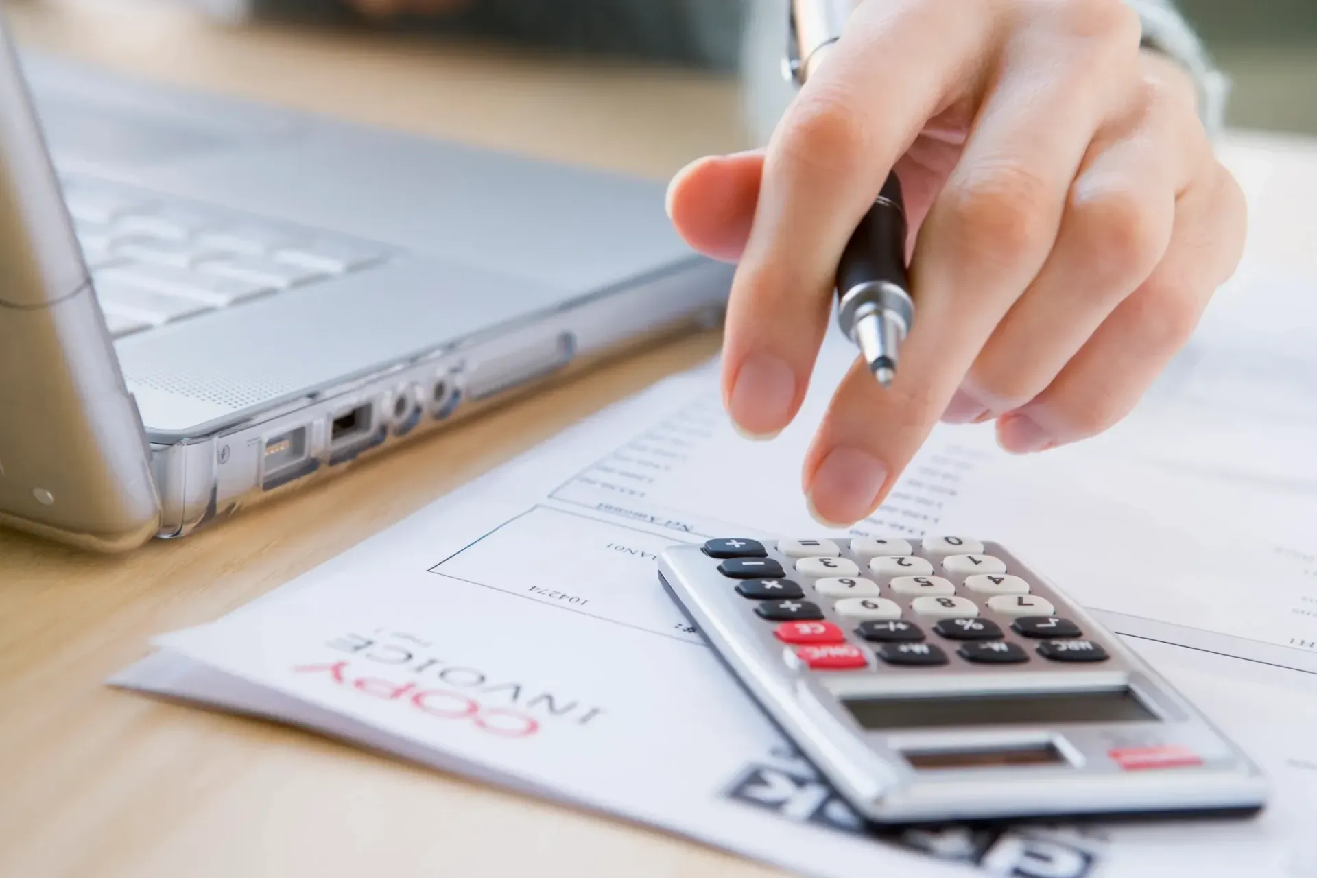 A person is using a pen and a calculator in front of a laptop.