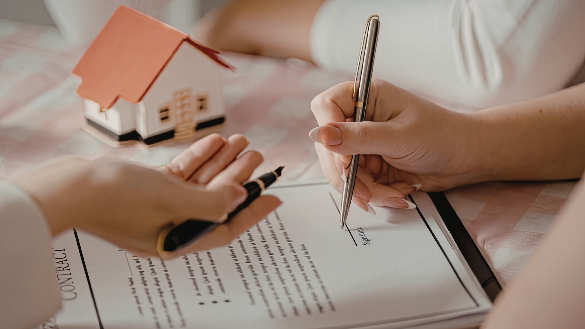 Two people reviewing a document near a small model house, one person holding a pen over the signature line.