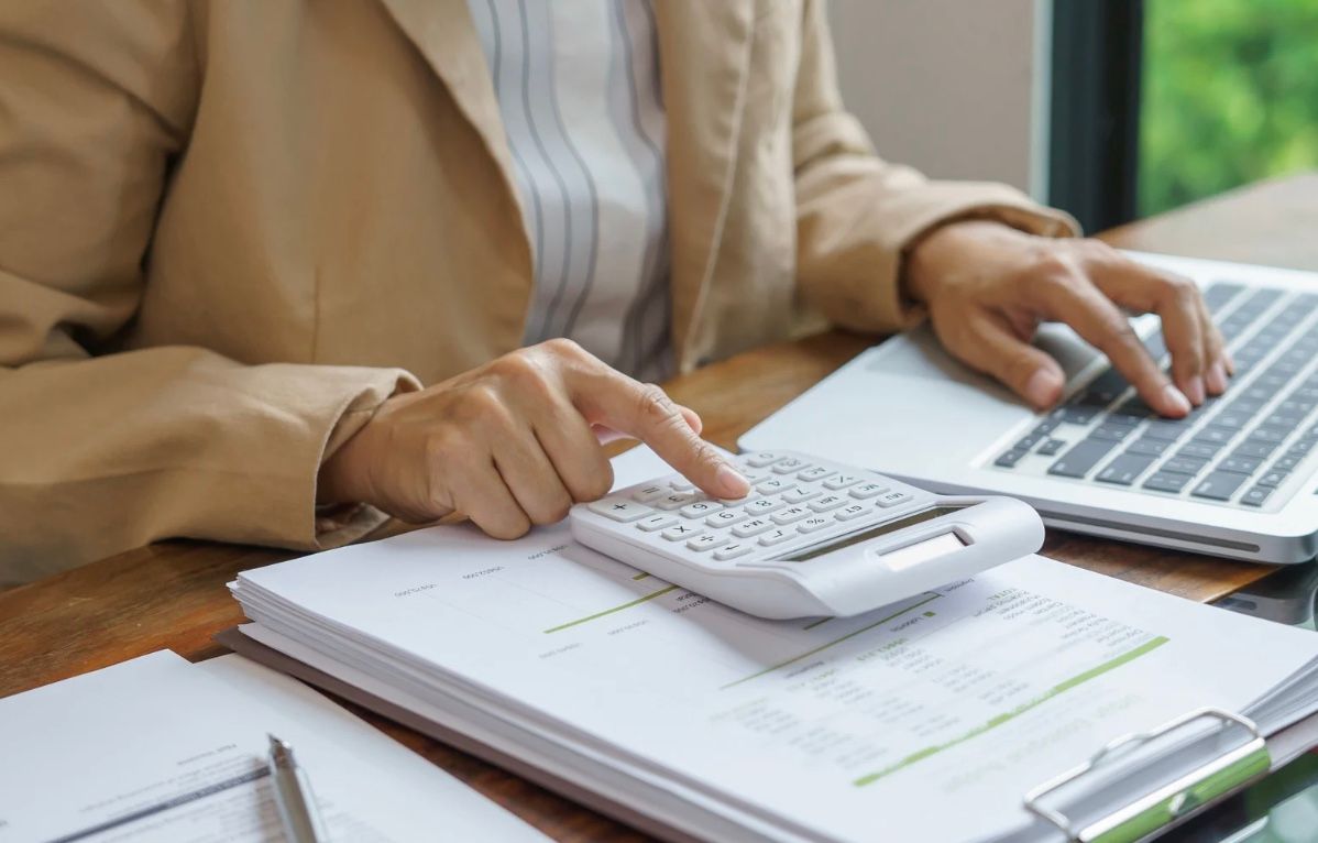 Hands using a calculator and laptop on a desk with papers and a pen organizer