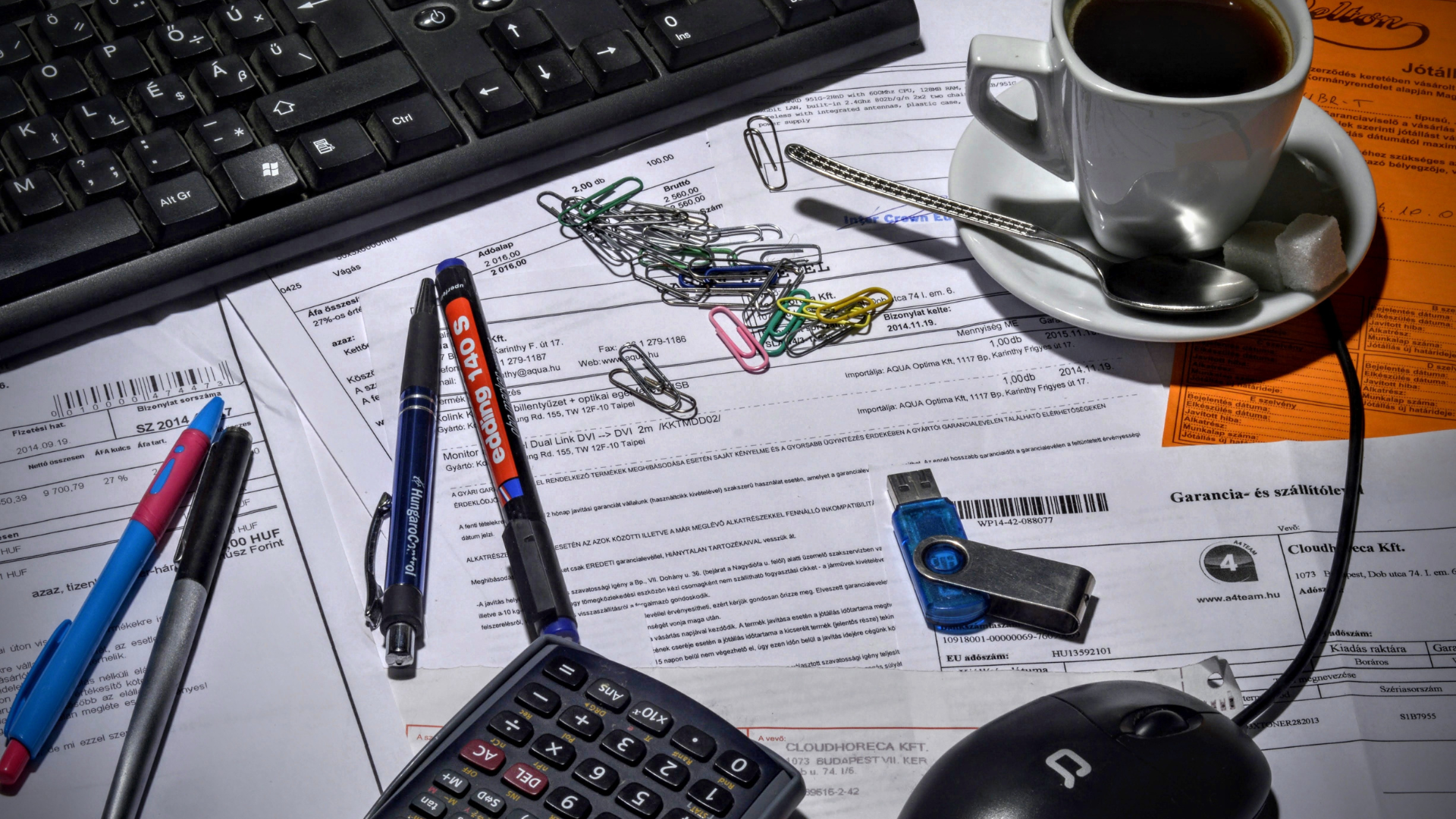 Desk with documents, keyboard, pens, calculator, USB drive, coffee, and mouse.