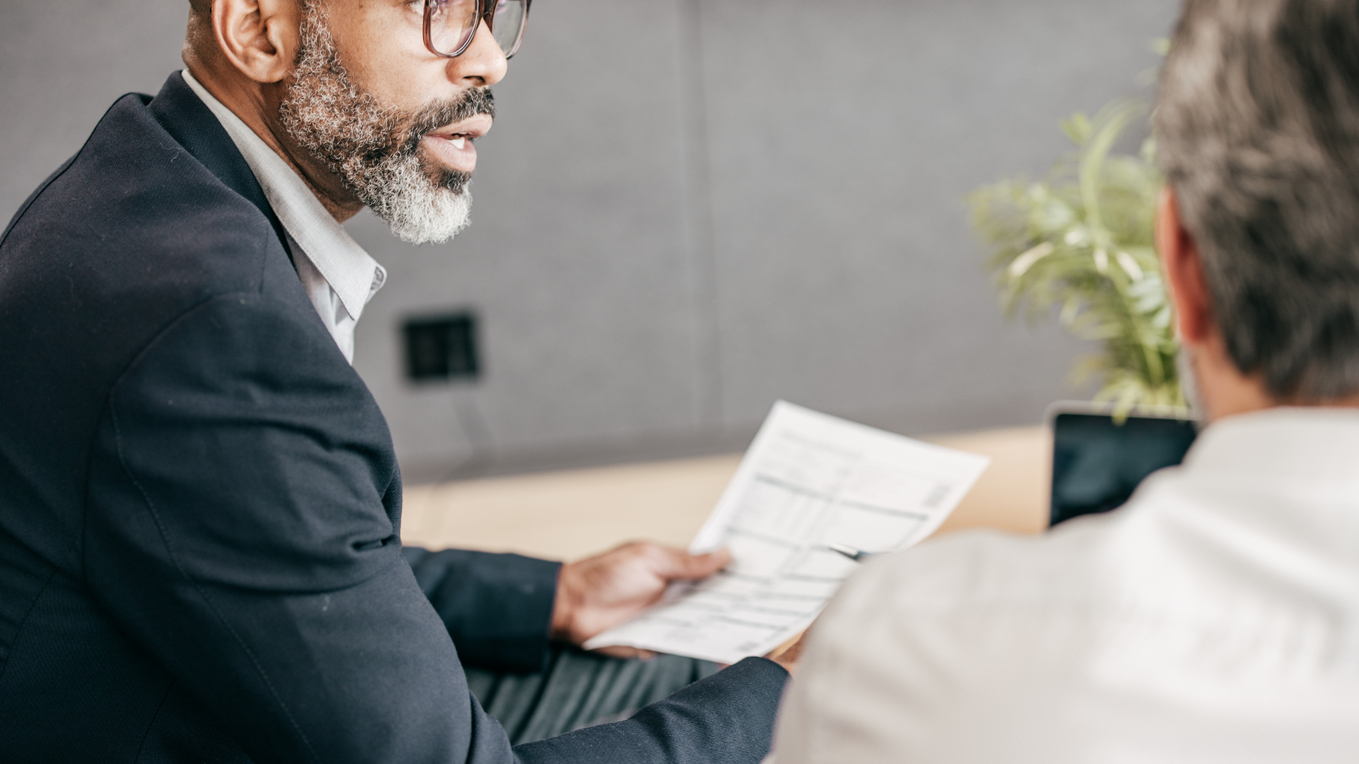 Man in glasses and suit gestures while holding paperwork, speaking to another person.