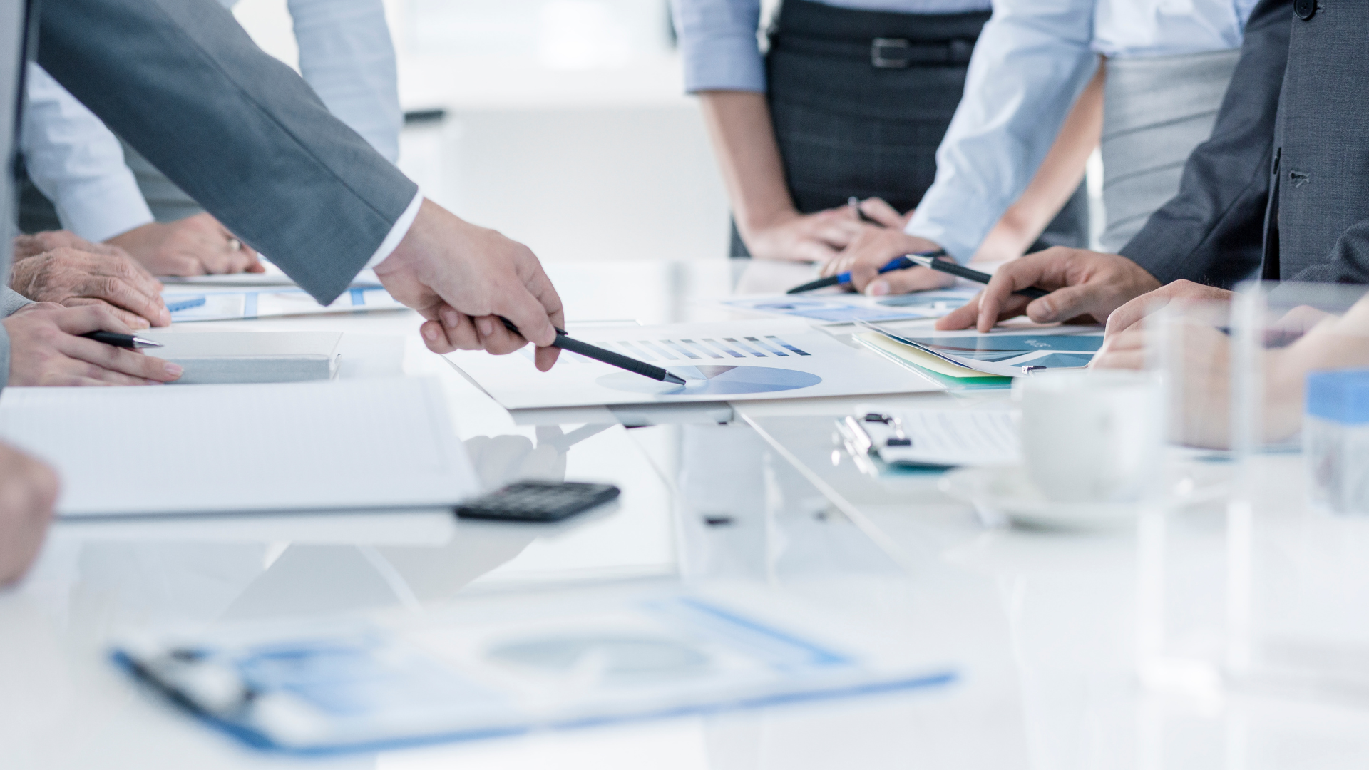 People in suits at a meeting, pointing at charts and documents on a white table.