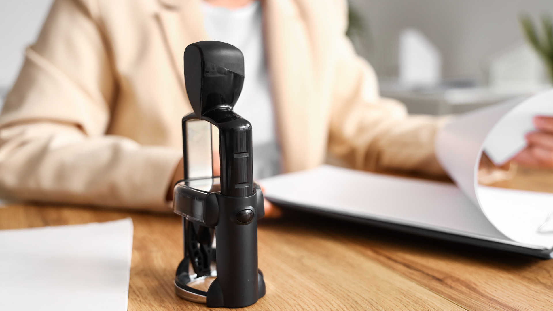 Black stamp on a desk, with a person in a beige blazer sorting documents in the background.