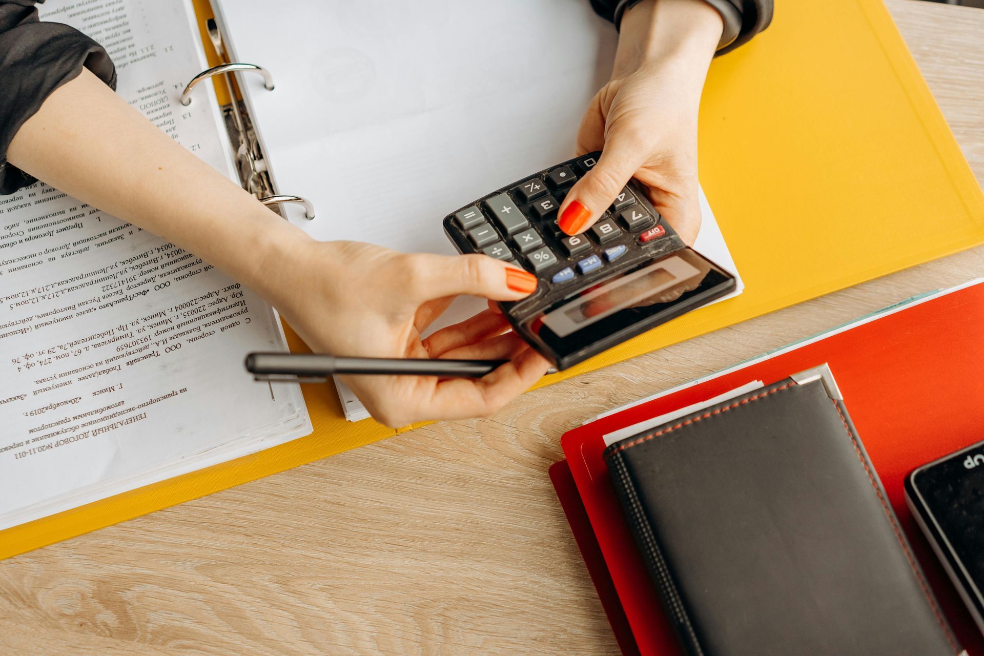 Hands using a calculator to calculate notes on a document, surrounded by folders and a notebook on a desk.