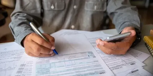 A man is writing on a piece of paper while holding a cell phone.