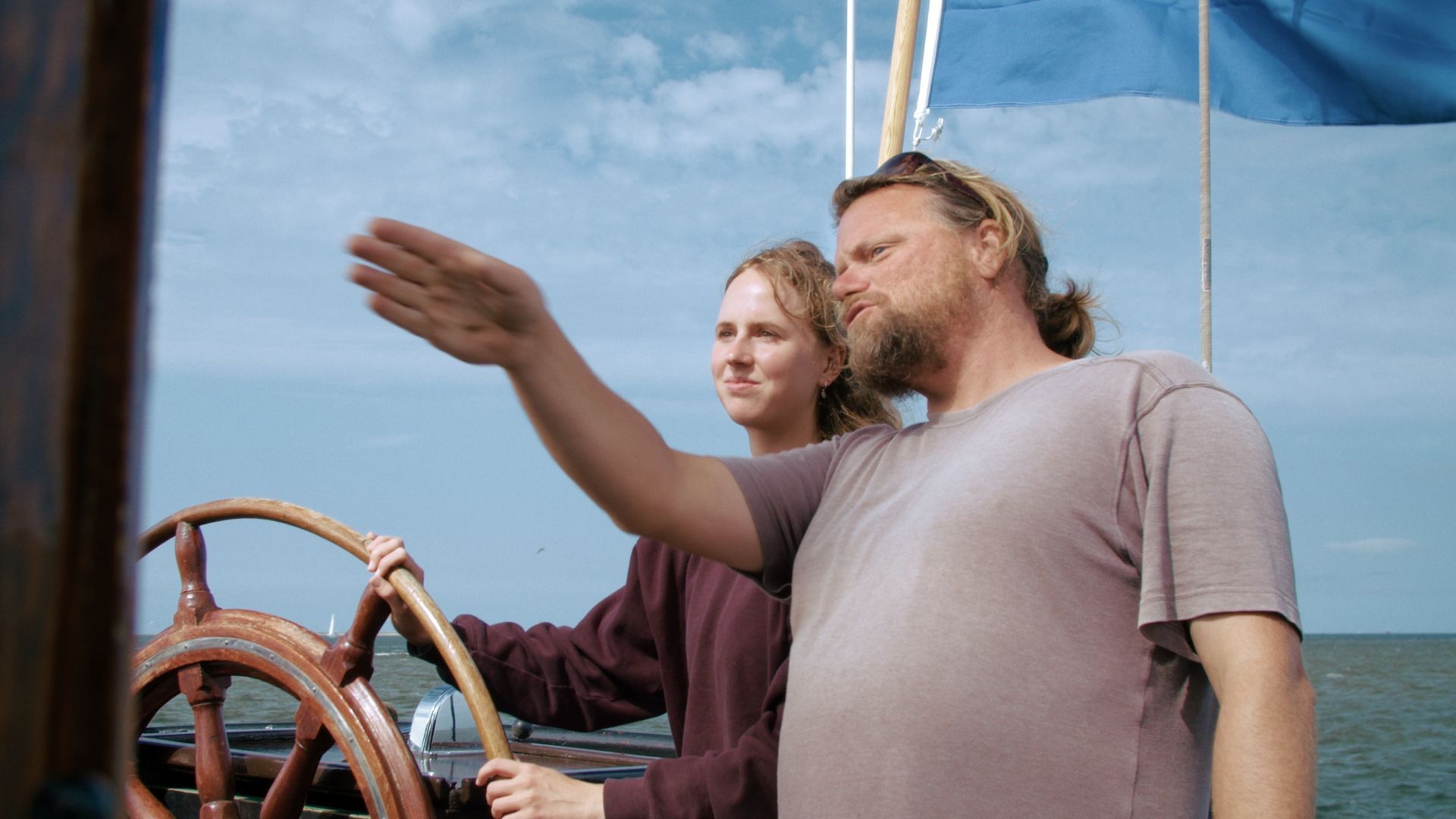 Mann zeigt zum Horizont, während er ein Segelschiff steuert; Frau steht neben ihm.
