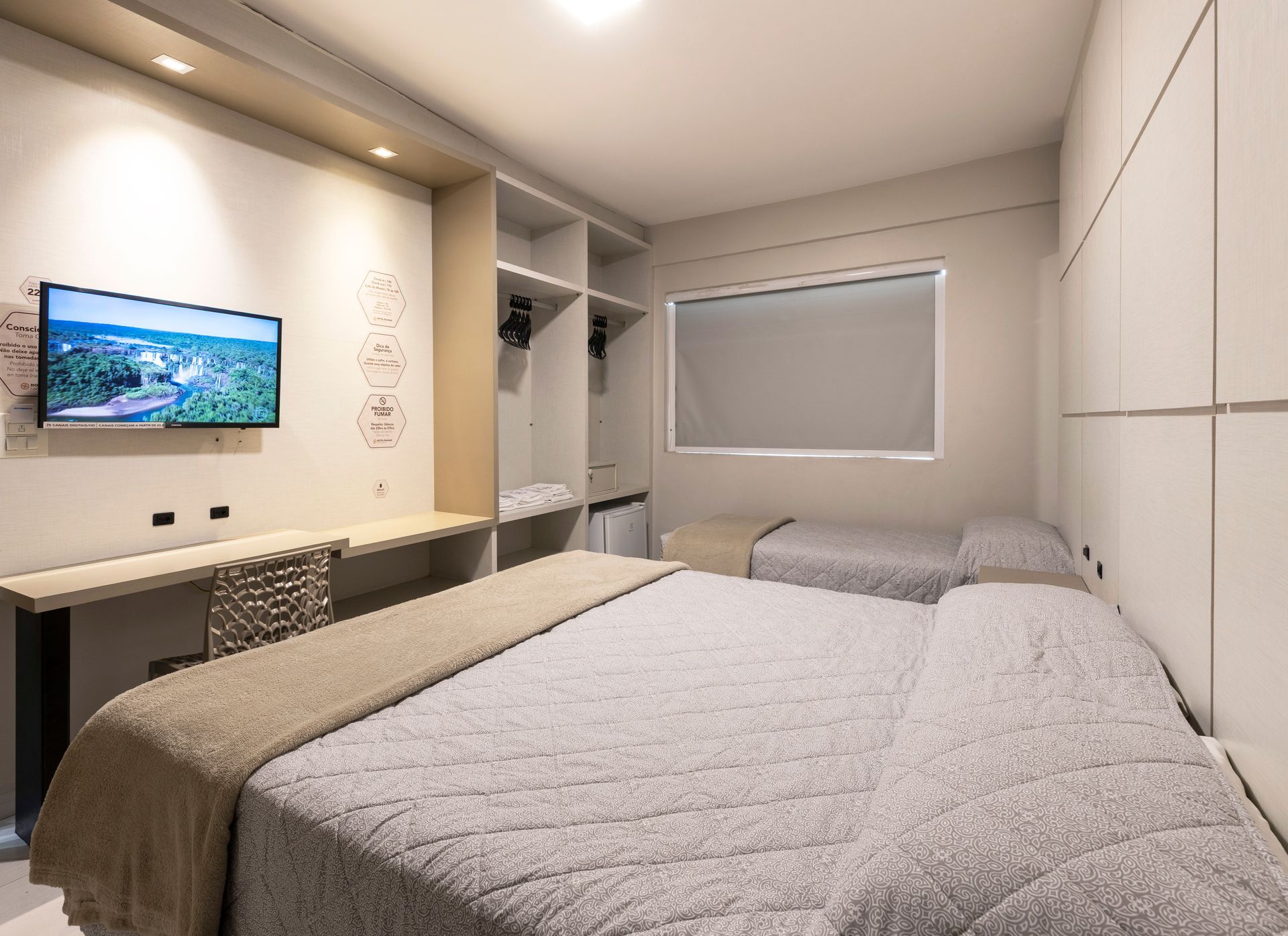 Quarto moderno com cama, TV de parede, escrivaninha, armário e janela com persianas.
