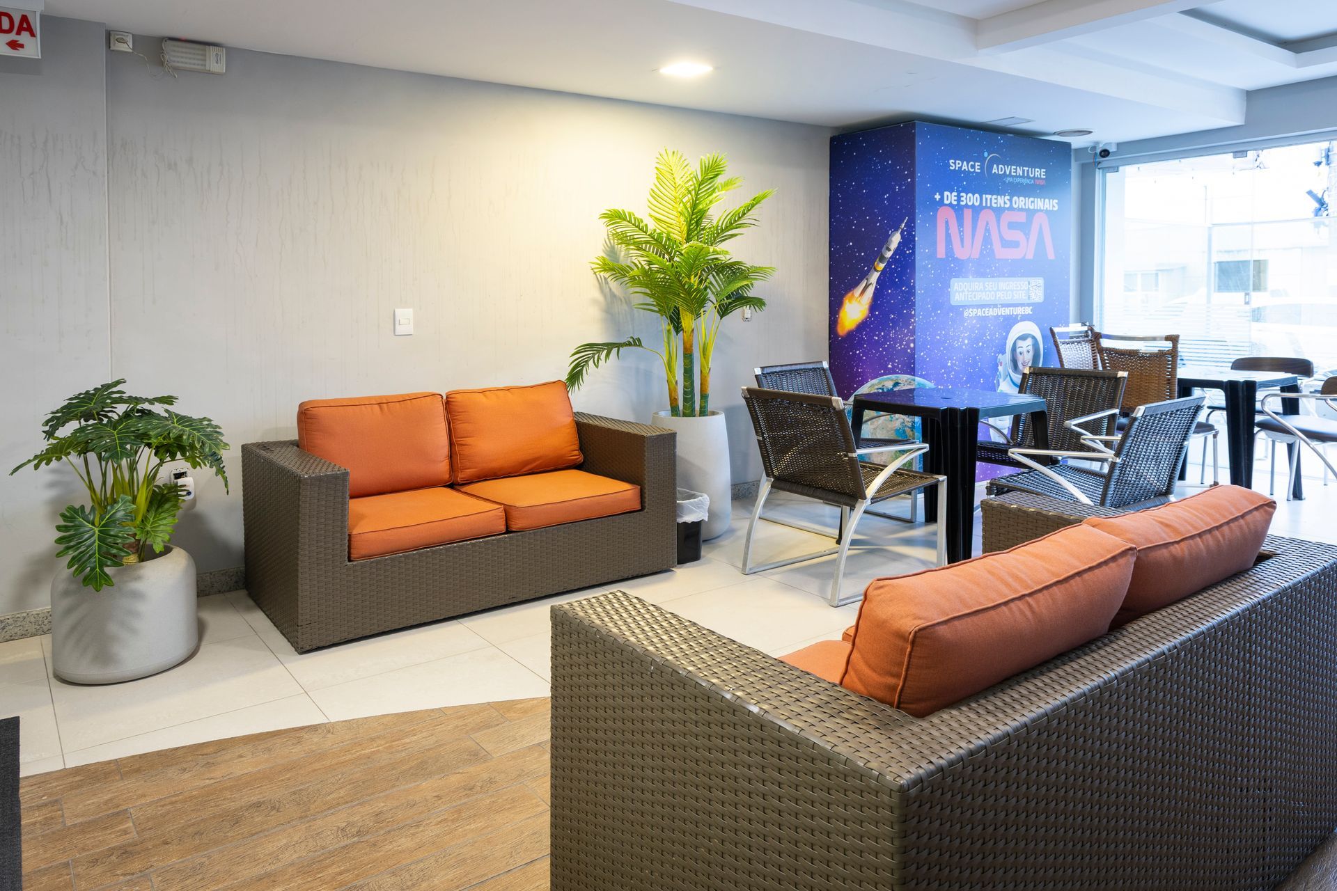 Sala de estar moderna com sofás laranja, plantas, mesa de vidro e banner da NASA ao fundo.