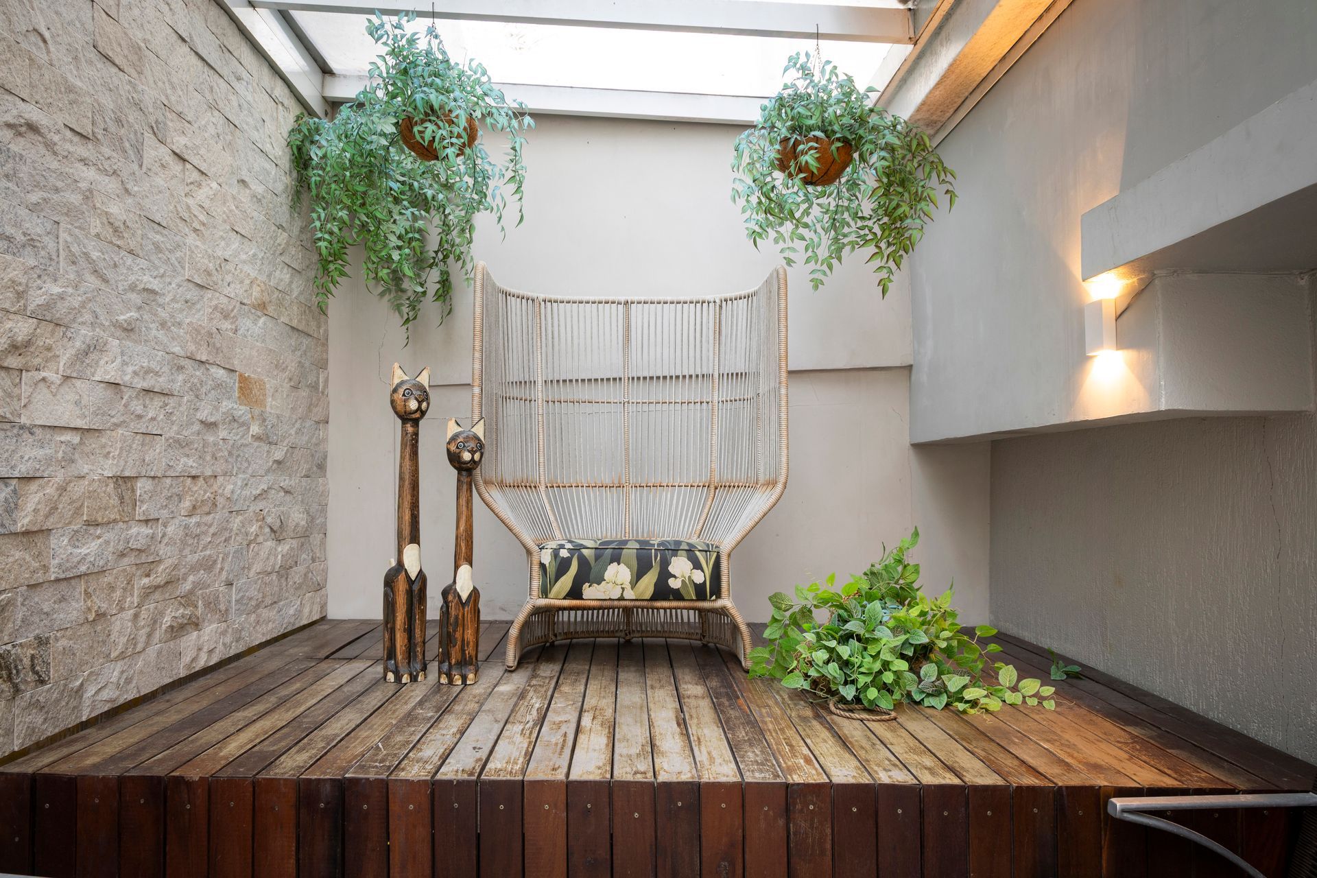 Deck de madeira com plantas suspensas, cadeira de vime e pequenas velas em um pátio interno iluminado.