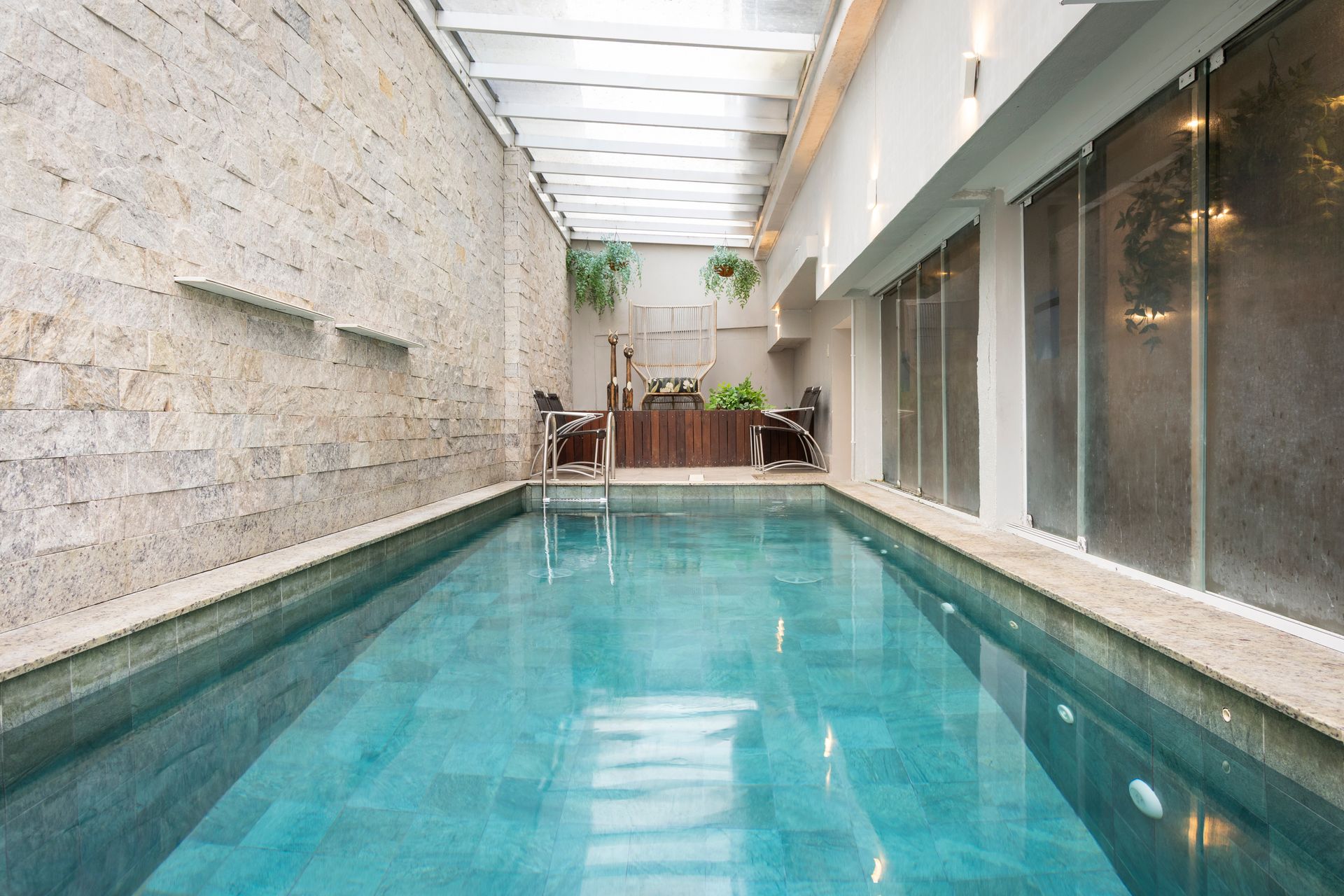 Piscina coberta para natação em um pátio estreito com teto de vidro, paredes de pedra e portas de vidro deslizantes.